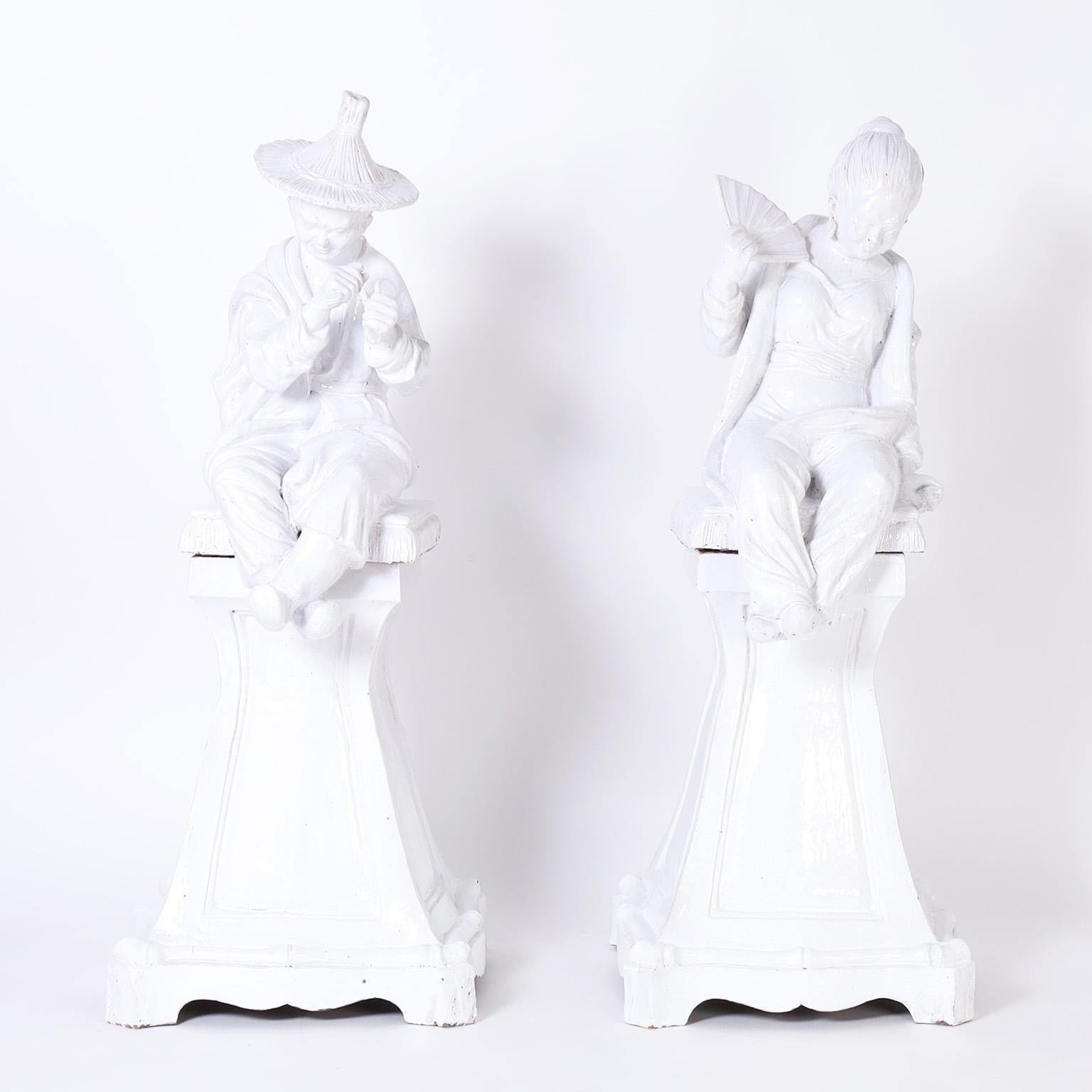 Striking pair of mid century chinoiserie figures crafted in terra cotta with a distinctive Italian white glaze, depicting a male and female figure seated on pillows over classical pedestals.