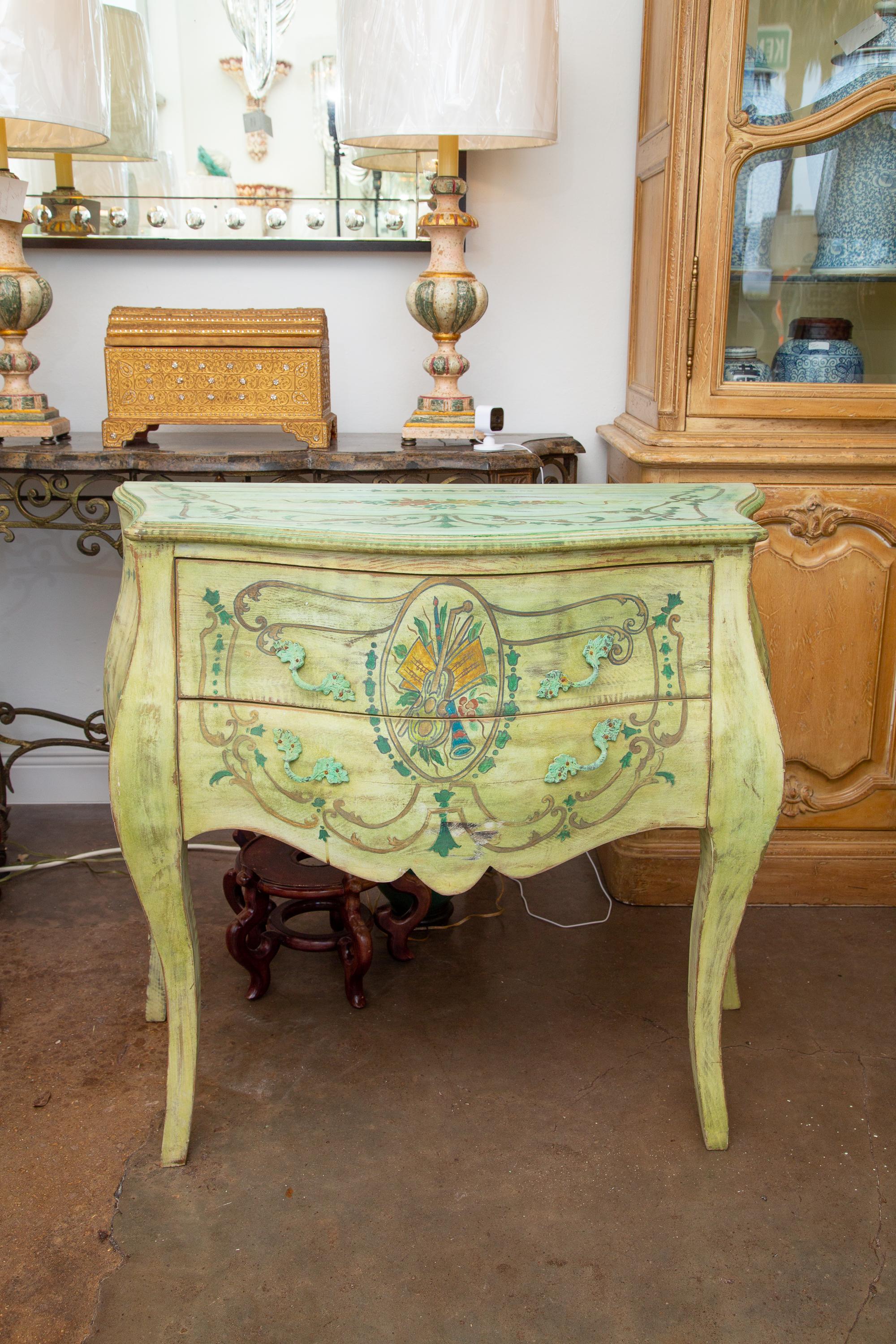This is a very decorative pair of Italian Rococo style commodes, painted light green over all, serving as background for a central figure depicting musical instruments, supported by cabriole legs, 20th century.