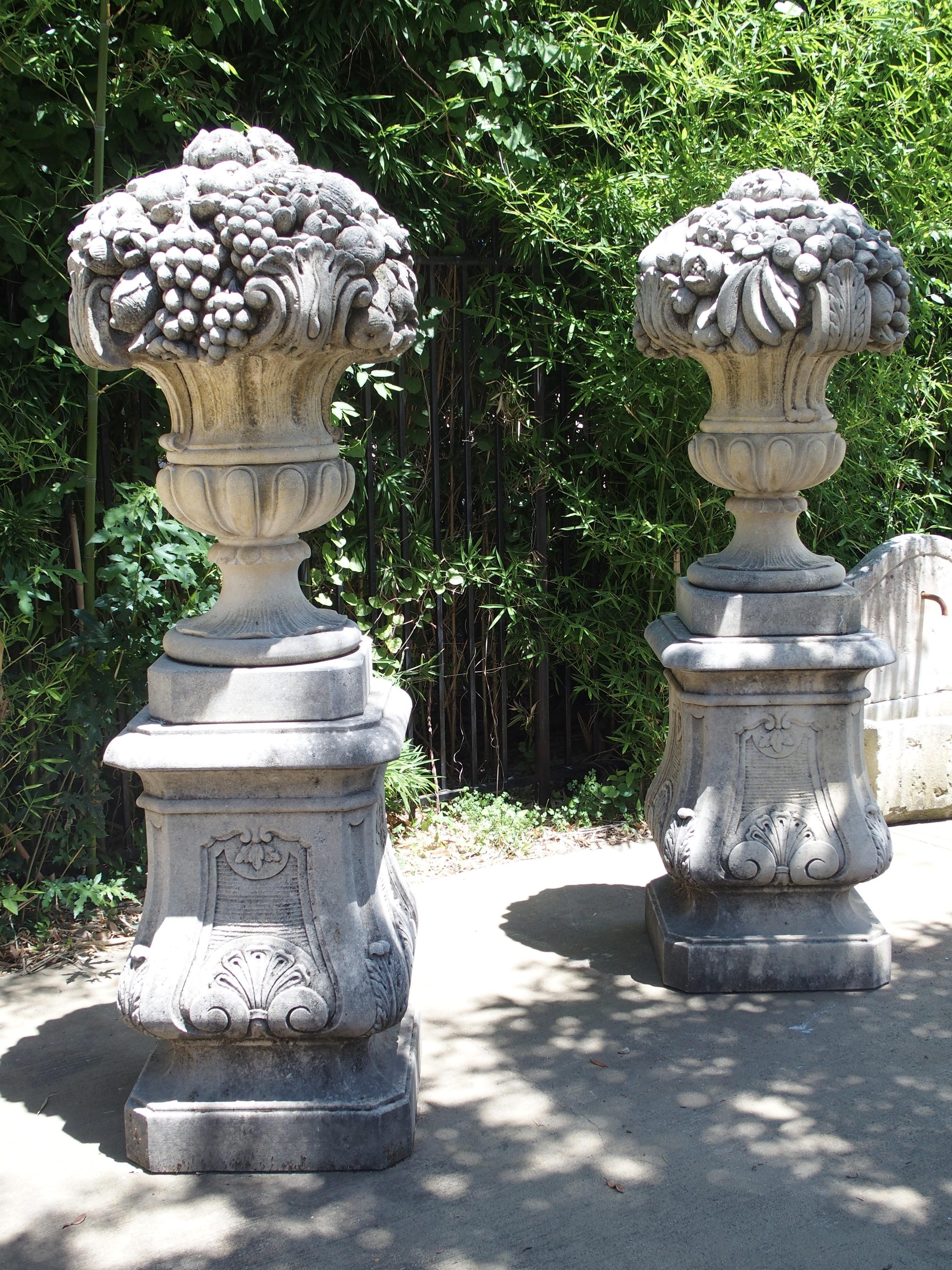 This is a set of large urns overflowing with fruit and flowers, standing upon matching bases. They are completely hand carved from a dense North Italian limestone. They are overflowing with flowers, bananas, grapes, pomegranates, and more. The urns