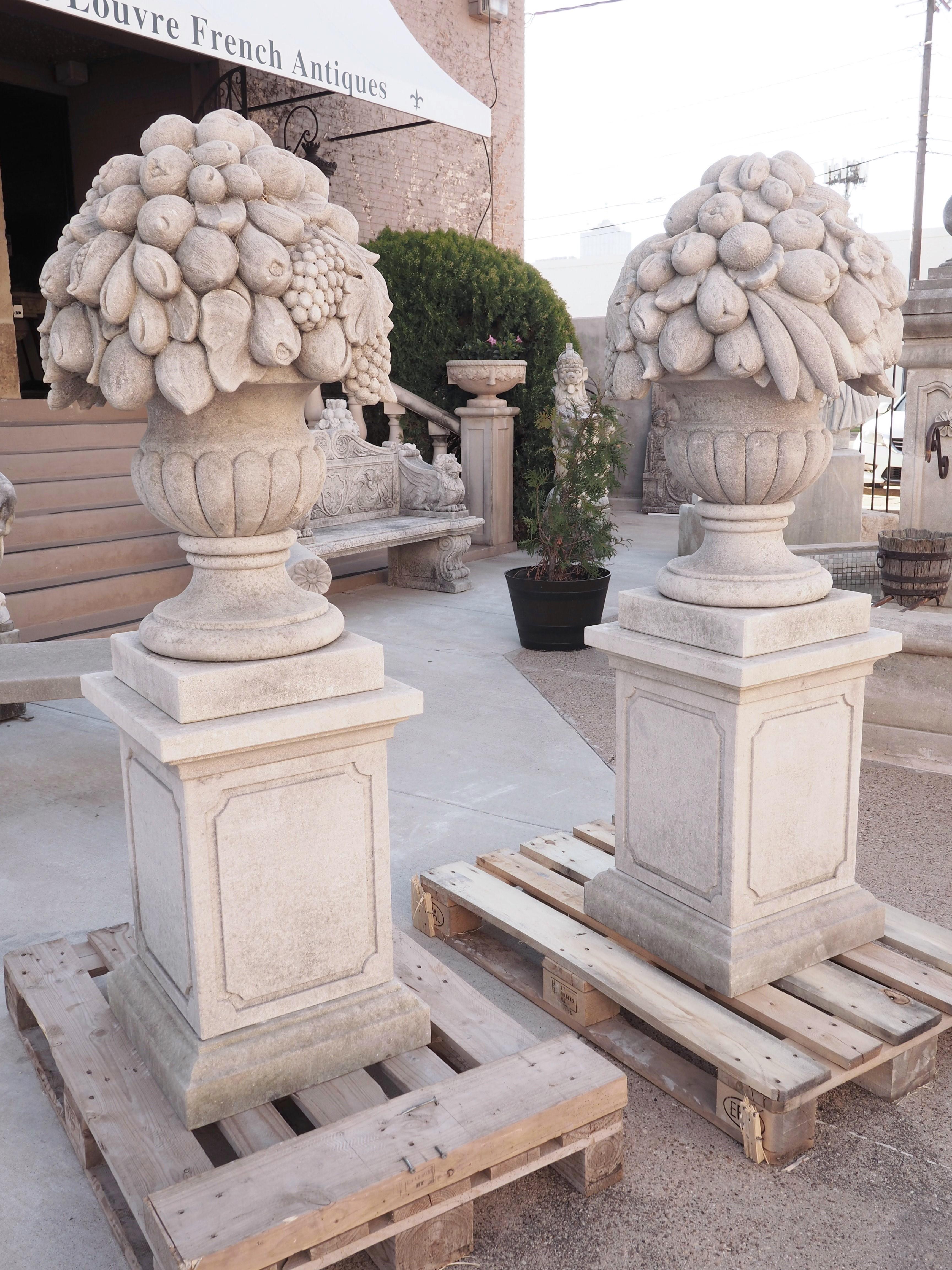 This impressive pair of fruit and floral bouquet urns on matching pedestals were hand-carved from Italian limestone. Both urns are overflowing with flowers, bananas, grapes, pomegranates, and more. The bulbous bodies of the campana shaped urns are