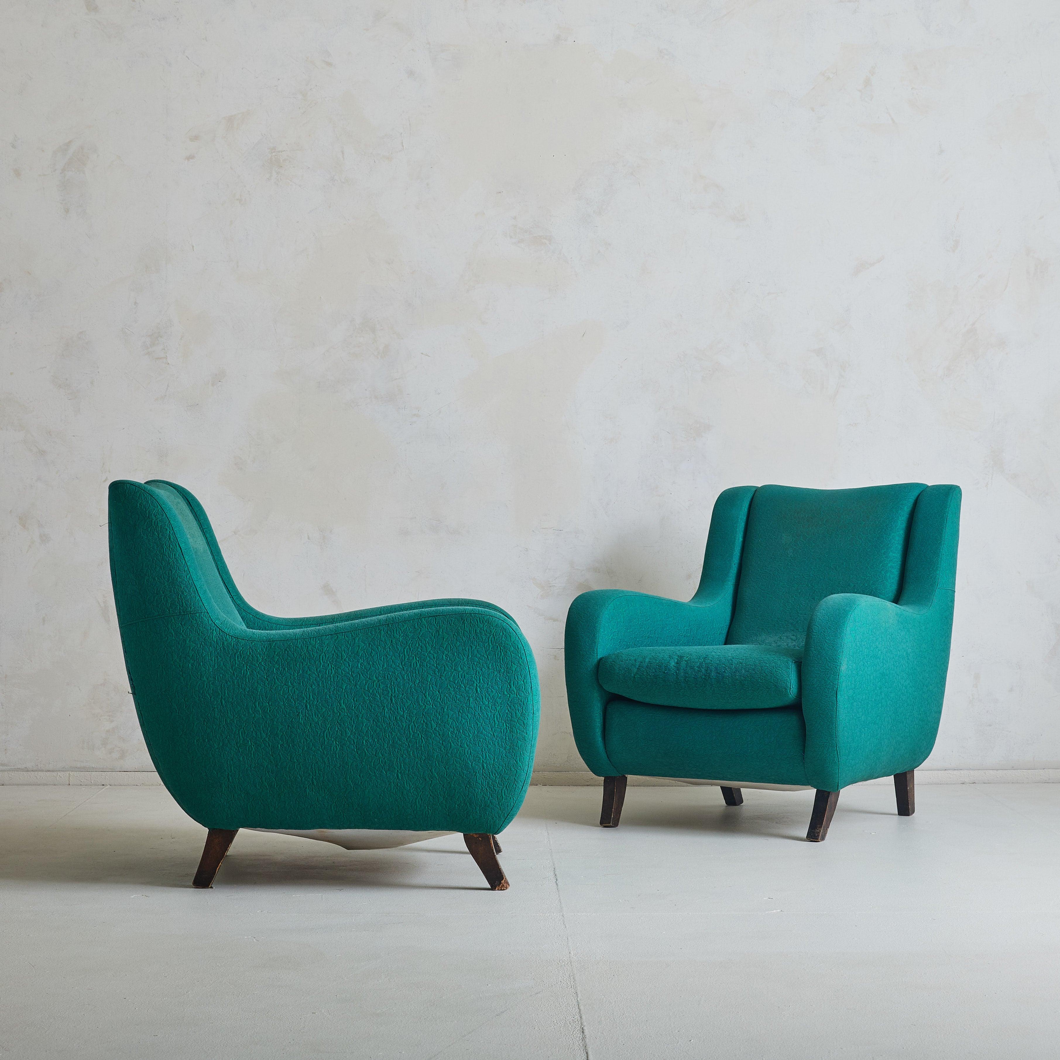 A pair of 1960s Italian lounge chairs, embodying bold curves and refined craftsmanship. These chairs feature sinuous, sculptural lines and are upholstered in original vibrant teal fabric. The gently contoured backrests and plush seat cushions offer