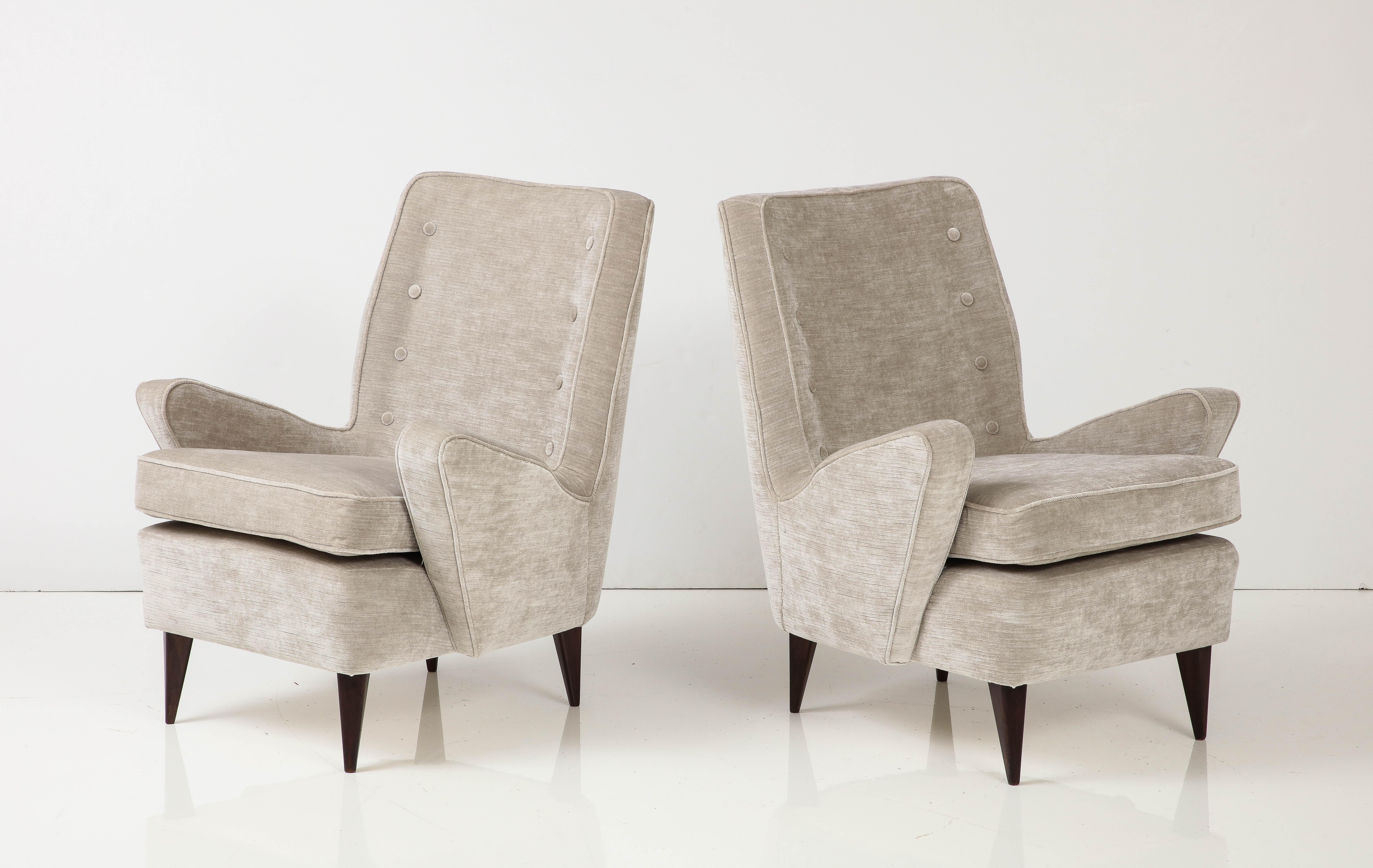 A pair of Italian midcentury lounge chairs featuring angled arms emanating from curved tight backs, each with eight tufted buttons and self welting, the seats with loose seat cushions resting on refinished wood legs newly, upholstered in greige