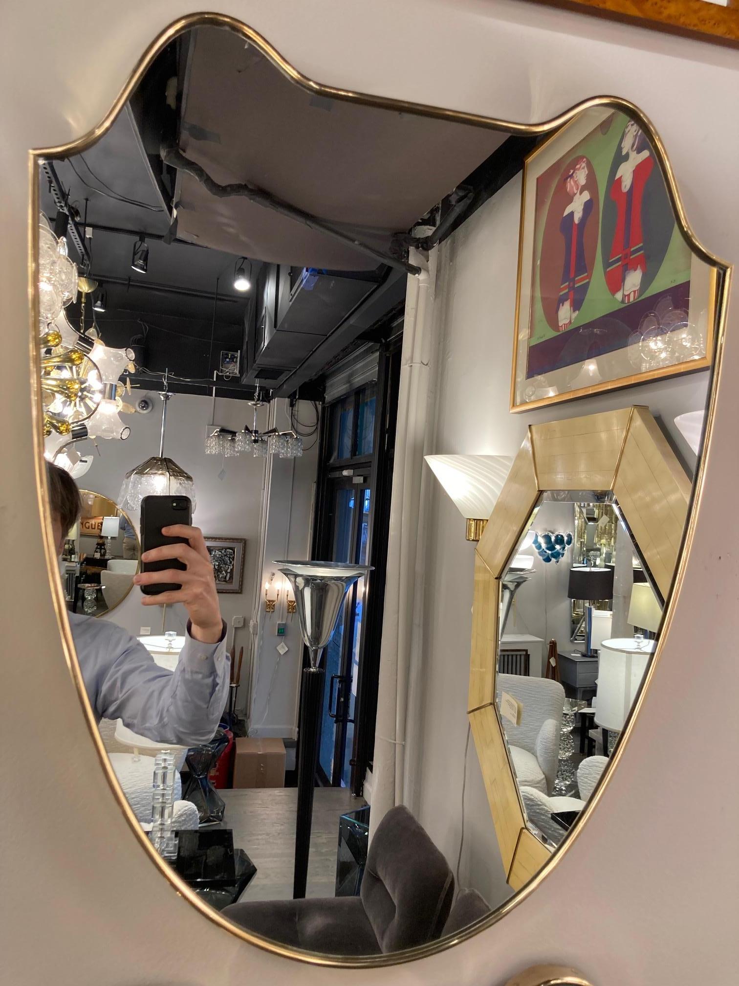 This stunning pair of Mid-Century Modern mirrors were realized in Italy, circa 1950. They feature amorphic sculptural bodies with two convex peaked forms at their apex wrapped in brass (with brass rivets) securing the frame and a clear mirror