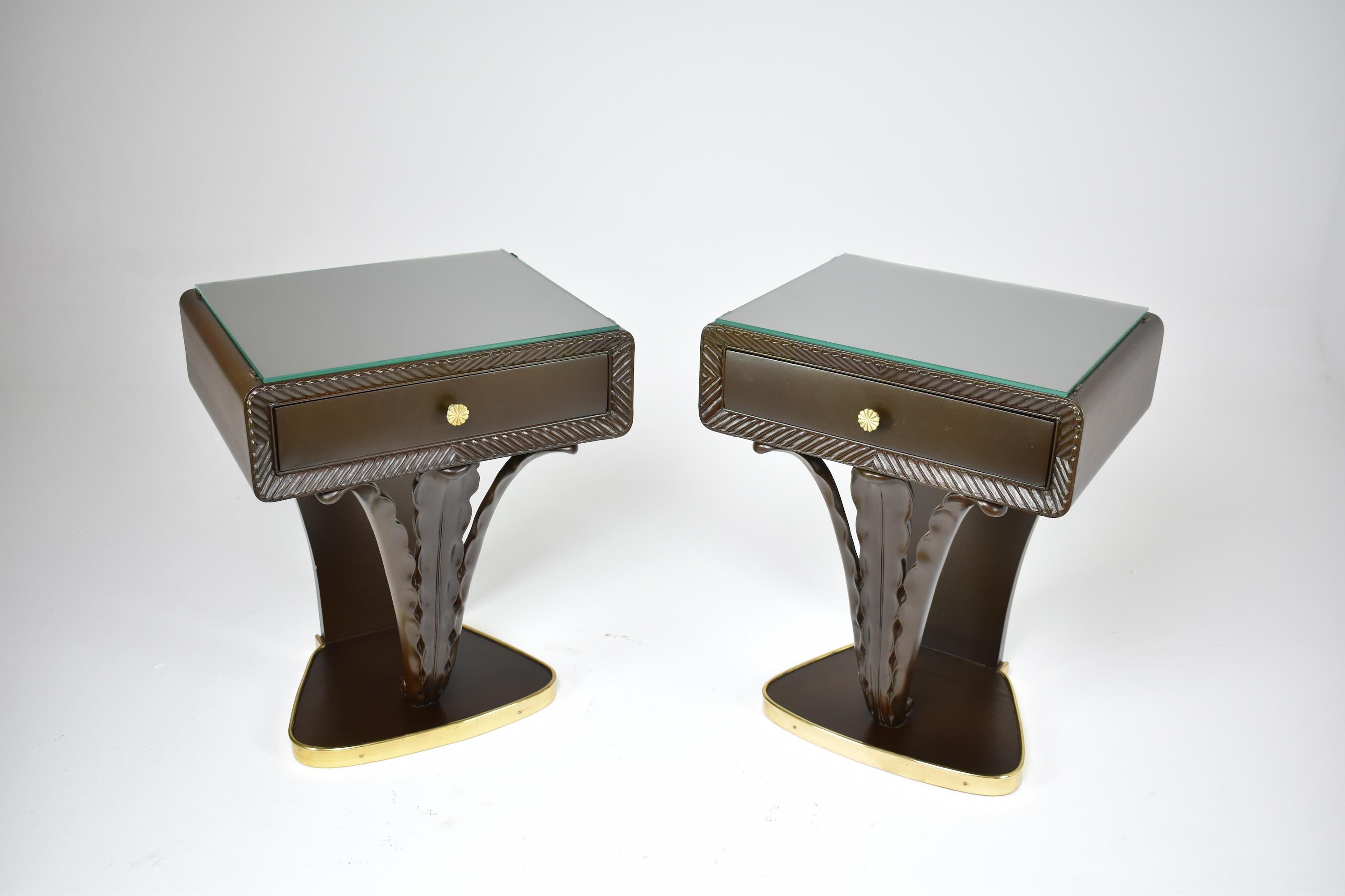 A very stylish pair of Italian mid-century nightstands crafted out of dark solid wood and beautified by a brass rim on the base, meticulously sculpted details and a clear glass tabletop. 
These 20th-century vintage side tables have a distinctive