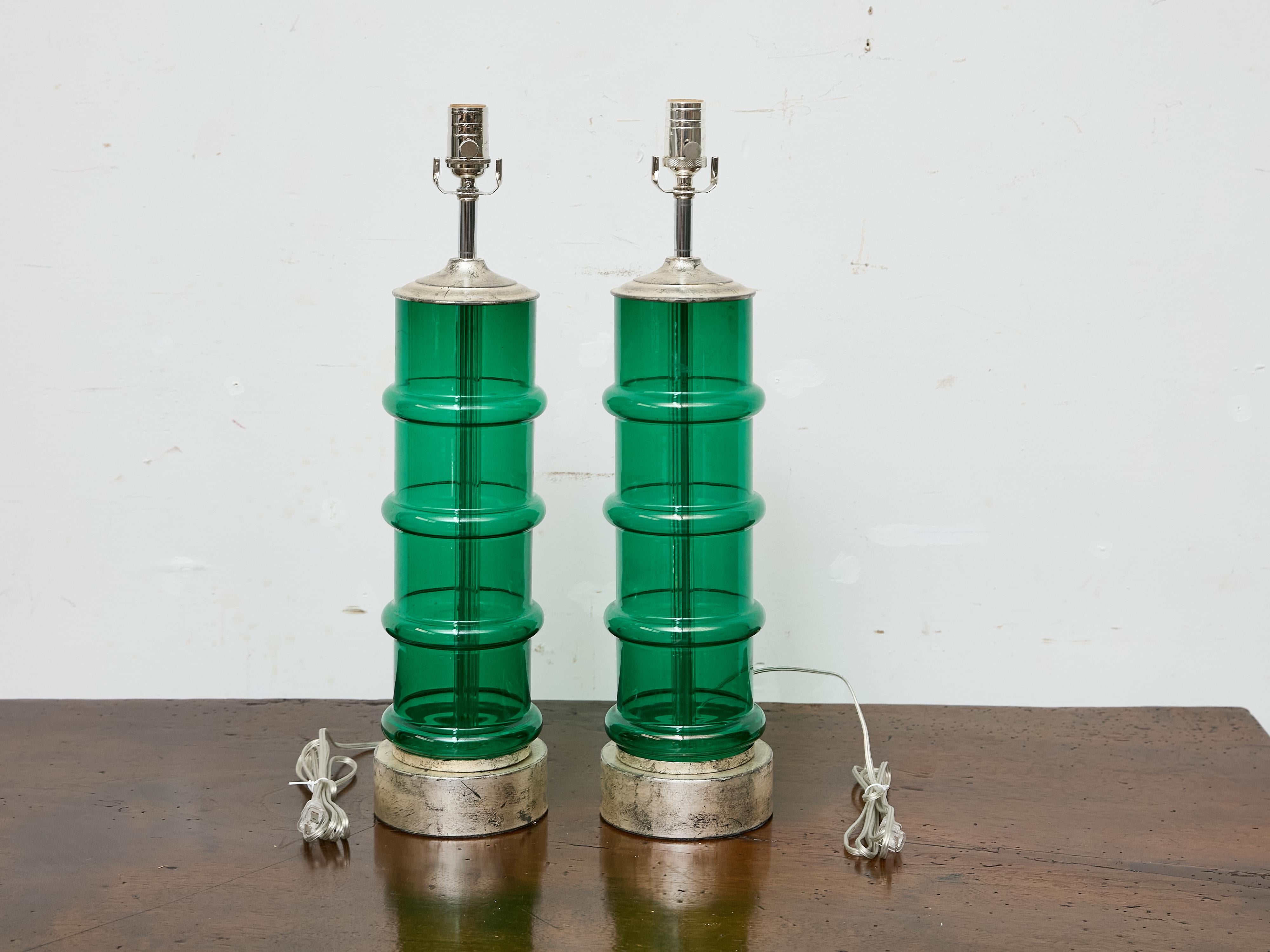 This striking pair of Italian Midcentury table lamps brings sculptural presence and vibrant color to any interior. Crafted from thick, ribbed green glass, the cylindrical forms rise from round silvered metal bases and are capped with matching metal
