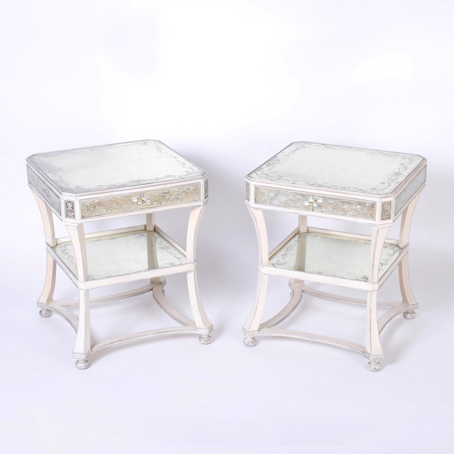 Pair of Italian two-tiered nightstands or end tables featuring reverse painted panels decorated with lovely floral designs and a neoclassic wood frame painted white with silver highlights over bun feet.
 