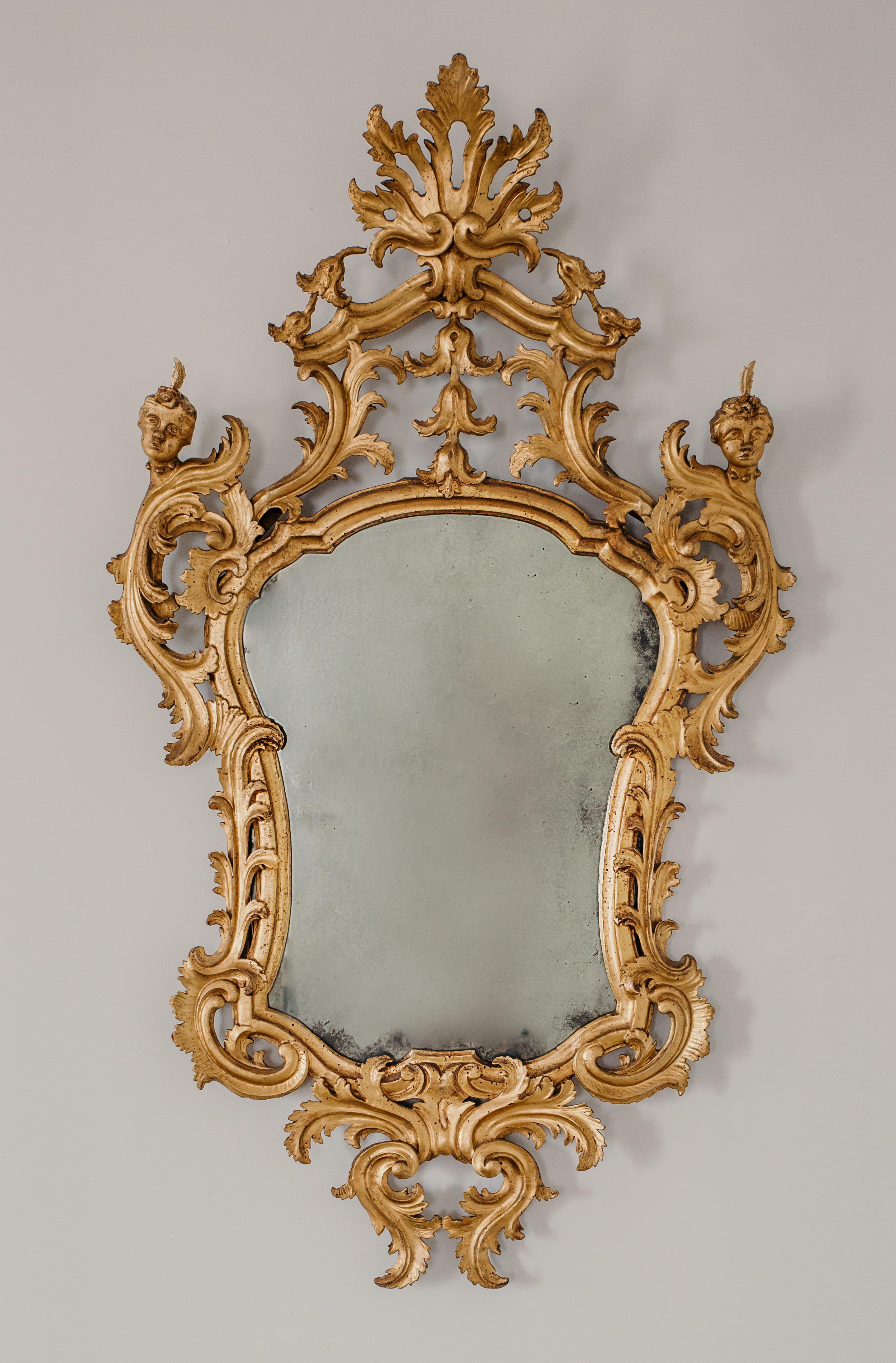 This beautiful pair of gilded mirrors was made in 18th century in Italy.
They have a gilded wooden frame carved with foliate motifs with a rich cymatium at the top. On the upper sides we find the presence of a rare pair of sculpted heads. A