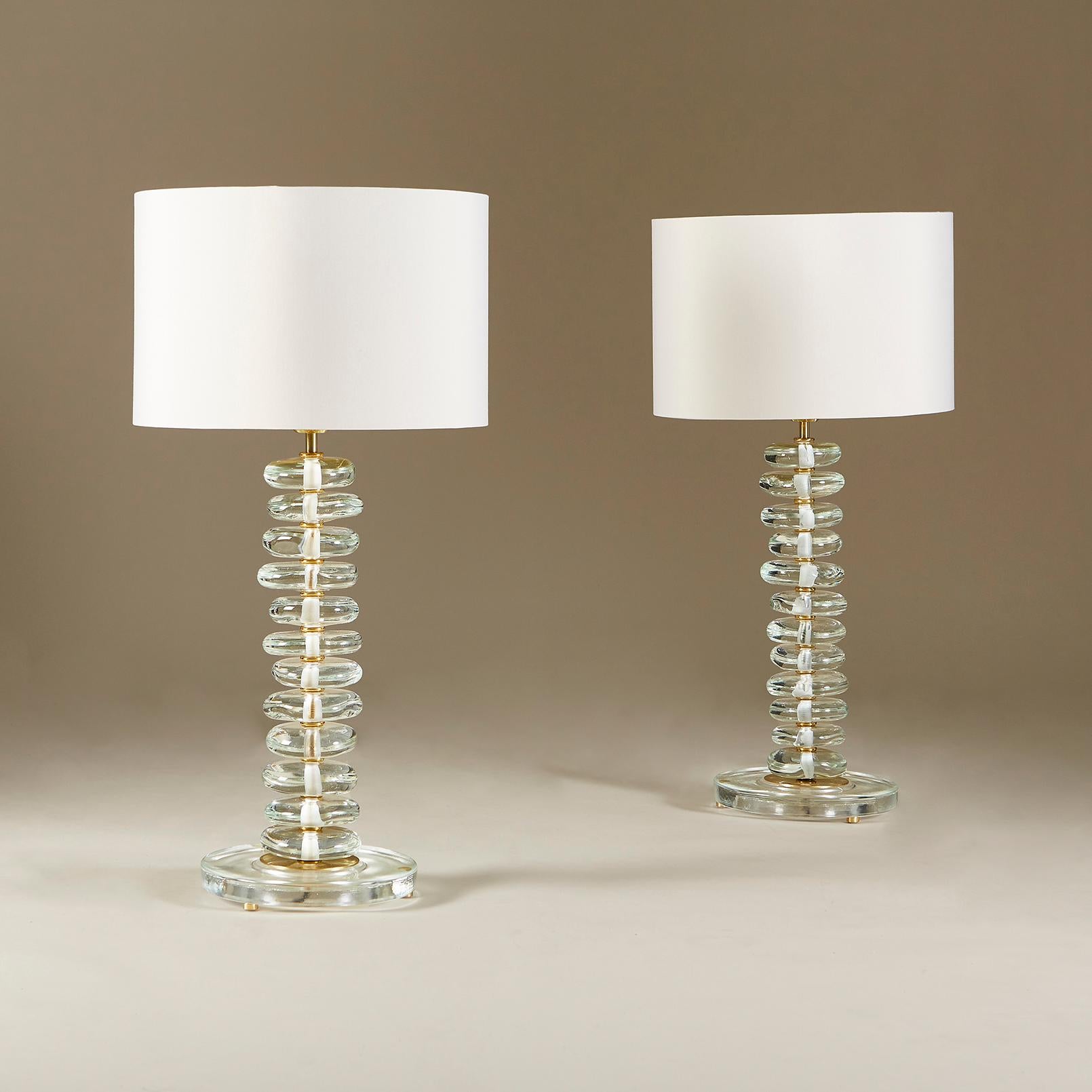 Tall pair of contemporary table lamps. Each lamps consists of 12 handmade smooth sculptured clear Murano pebbles. Each pebble is interspersed with a layer of brass. Sits on circular glass base with brass detailing and feet. The overall effect is