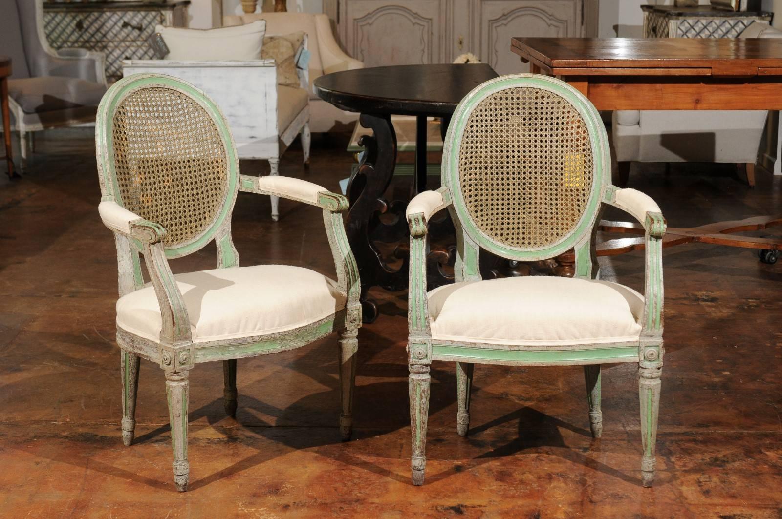 A pair of Italian neoclassical style green painted wooden armchairs from the mid-19th century, with cane oval backs and new upholstery. Each of this pair of Italian armchairs features a slightly slanted cane oval back, flanked with two partially