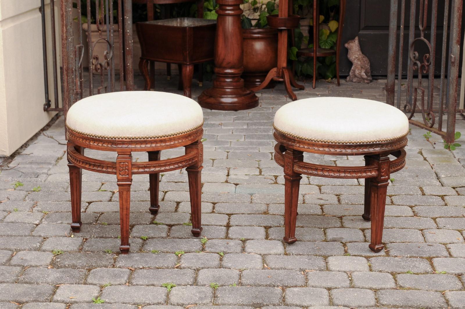 A pair of Italian neoclassical style fruitwood stools from the early 20th century, with tapered legs, carved apron, circular side stretchers and new upholstery. Born in Italy during the early years of the 20th century, each of this exquisite pair of