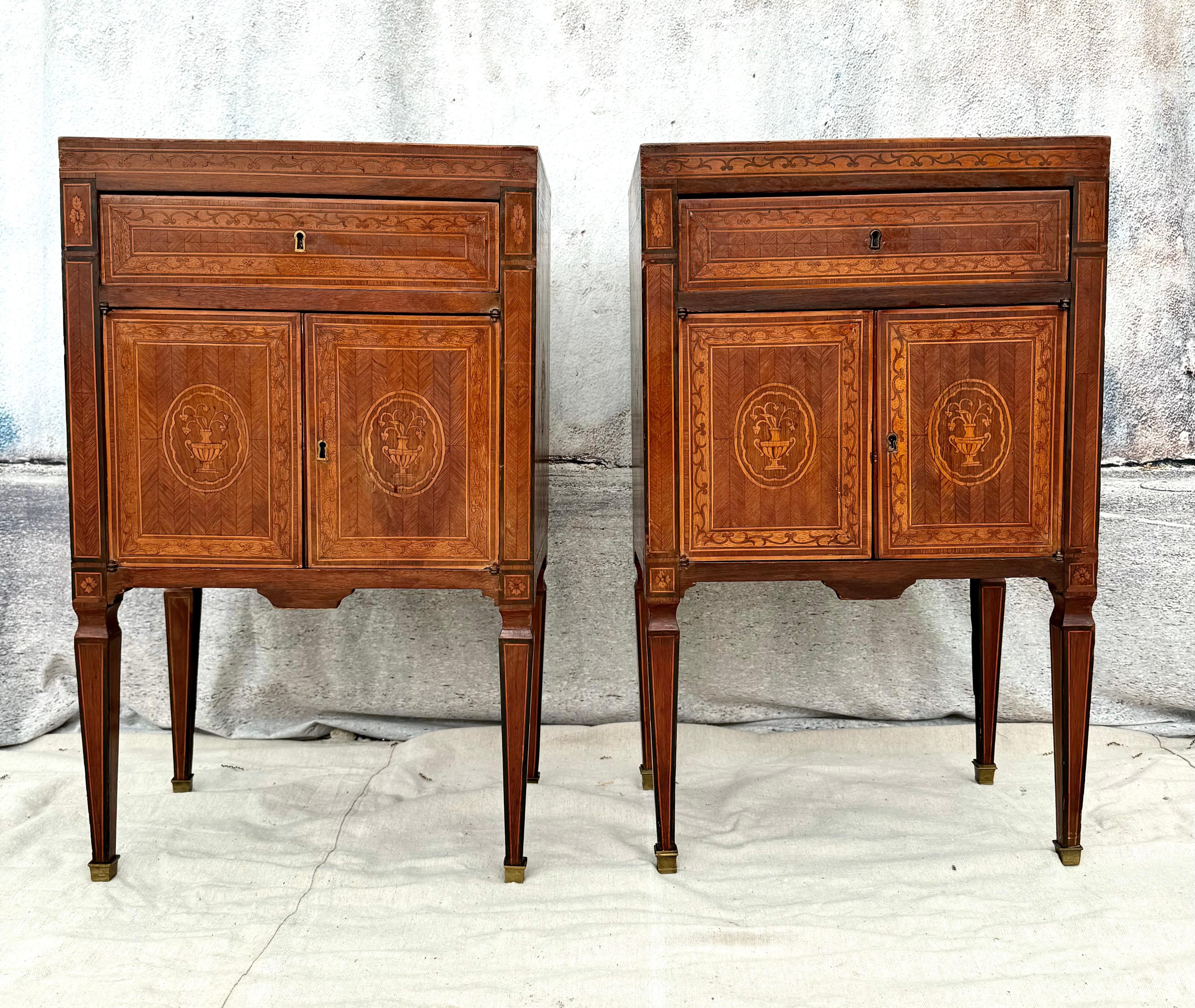 A pair of early 20th century Italian neoclassical style marquetry inlaid bedside cabinets. Each features one drawer over double doors that open to storage space. Cabinets have beautiful inlay of urns filled with floral arrangements on top and front