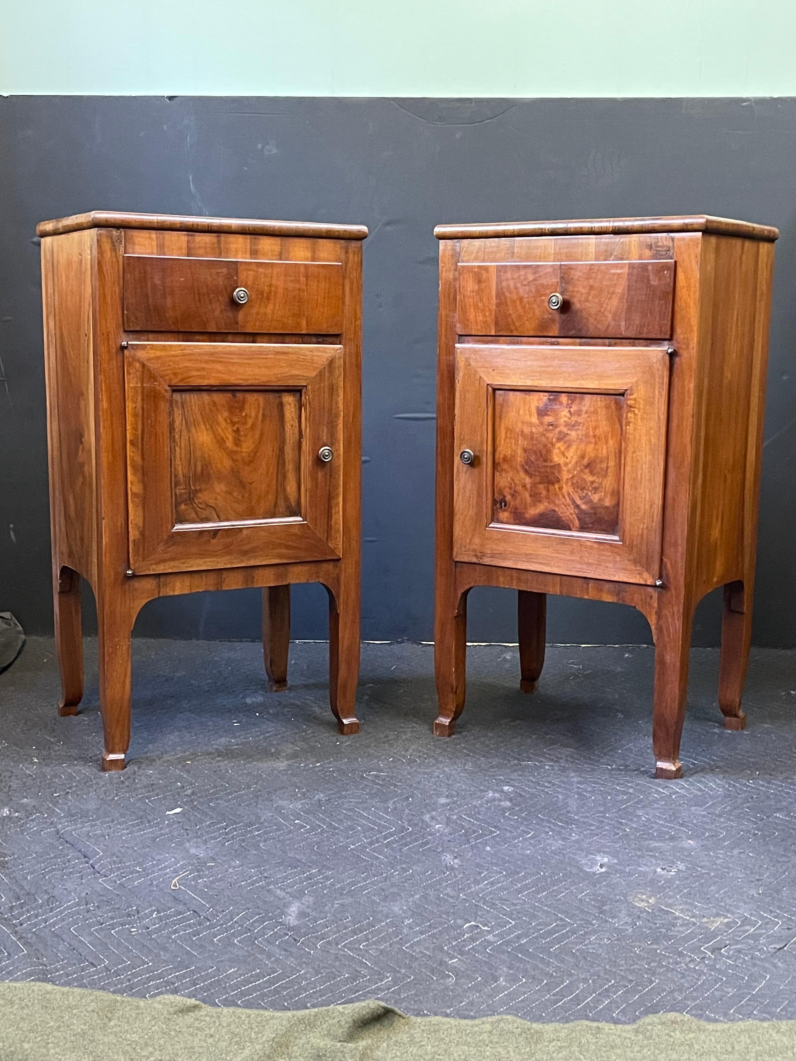 A 20th Century Italian Neoclassical facing pair of side cabinets or nightstands constructed of walnut. Each antique cabinet has a wood top with rounded edges, a classic case designed with clean lines, and a drawer over an enclosed cabinet with a
