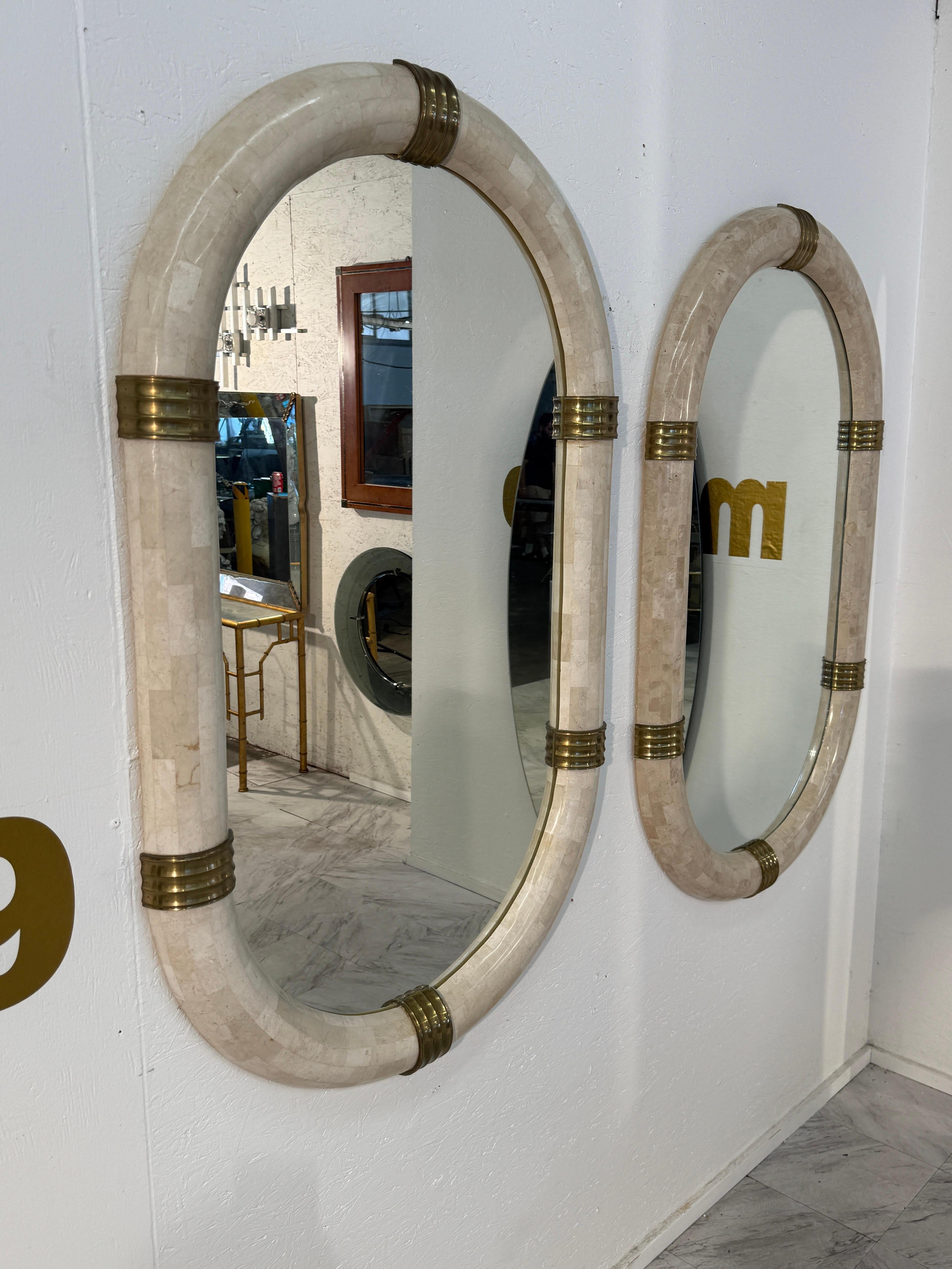 Pair of oval mirror crafted in Italy during the 1980s. The bold frame is made of tessellated stone in soft neutral tones, giving it a rich, textured appearance. Brass bands encircle the frame at four points, adding warmth and a refined contrast to