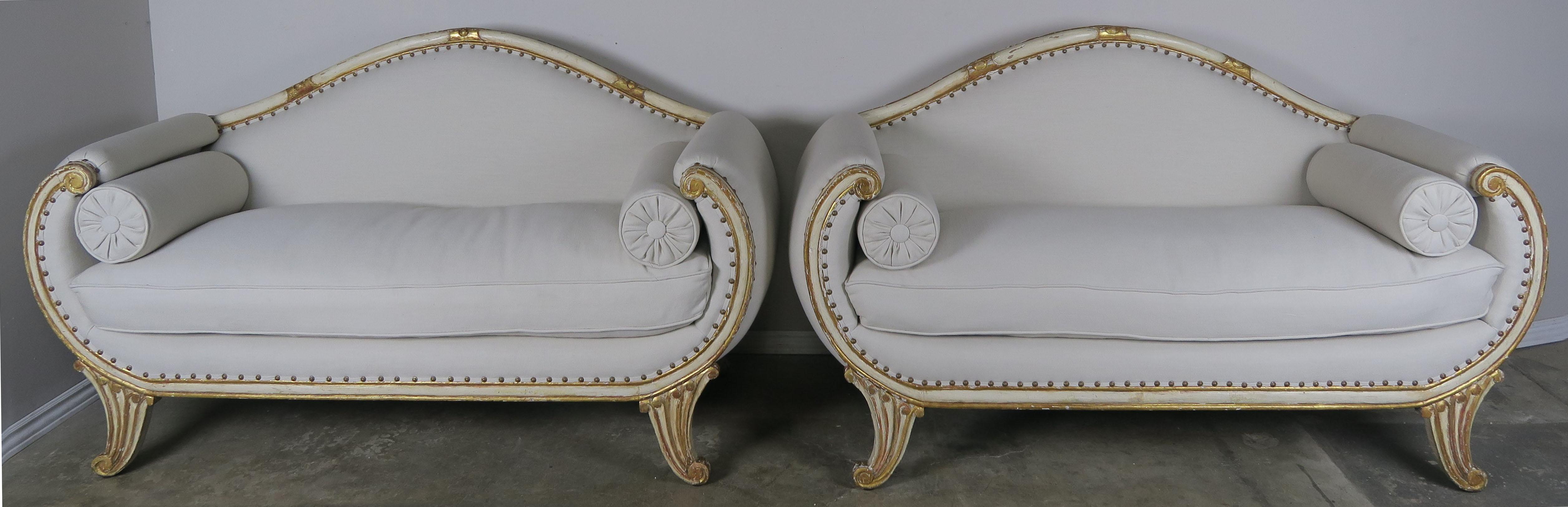 Pair of matching Italian cream painted and parcel 22-karat gold leaf settees with rolled arms and carved feet. The settees are newly upholstered in a Pindler & Pindler 100% linen textile with original nailhead trim detail. Loose down filled