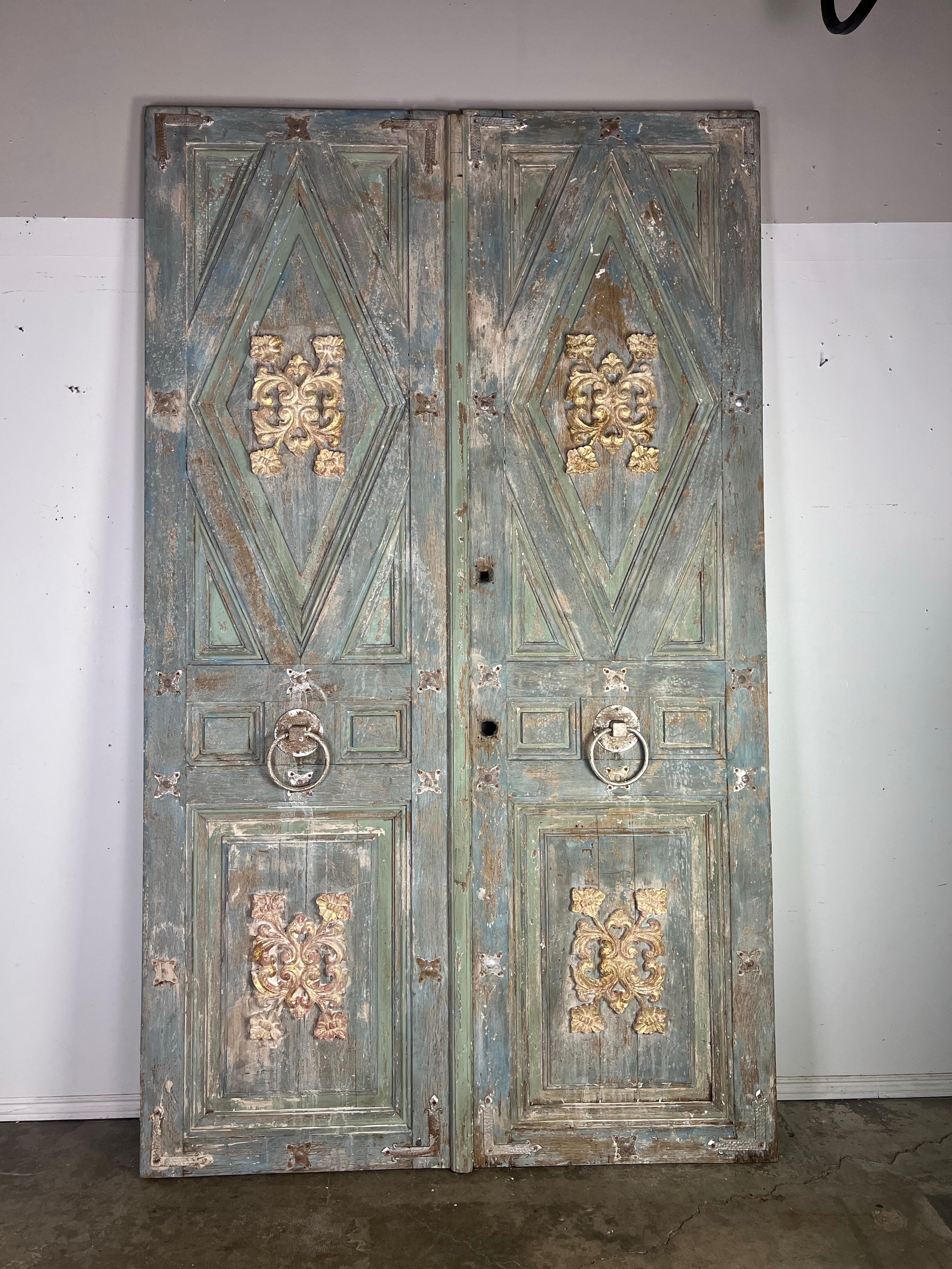 Coppia di porte doppie alte e rustiche con una finitura ornata e affilata.  Le porte sono dipinte in una tonalità verde-blu sbiadita, con il legno rovinato dagli agenti atmosferici e aree scheggiate che rivelano accenni del legno naturale
