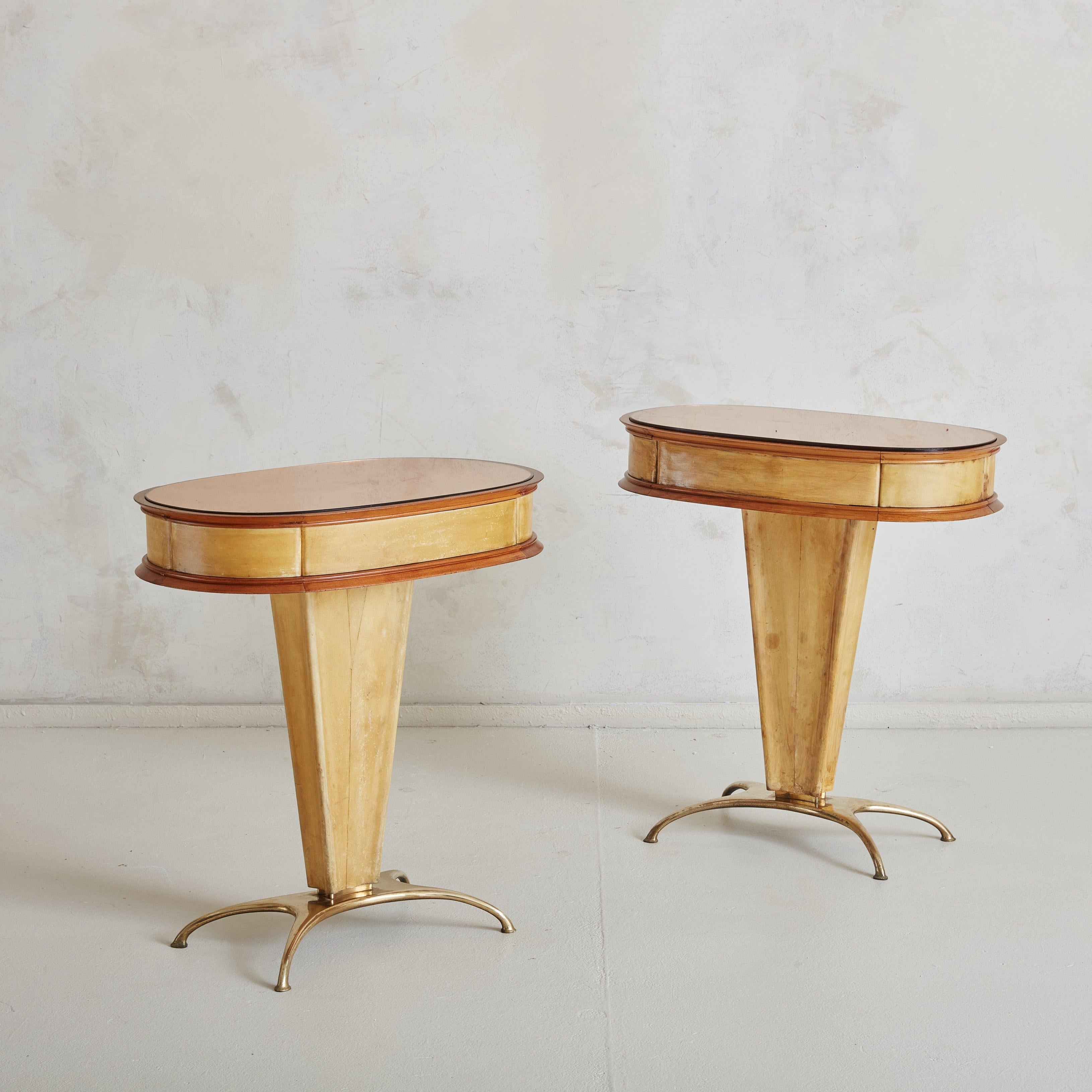 A pair of 1950s accent tables or nightstands attributed to Italian architect and designer Paolo Buffa, featuring parchment-wrapped surfaces and brass accents. Oval, rose-tinted mirrored tops rest above single drawers with scalloped brass pulls. The