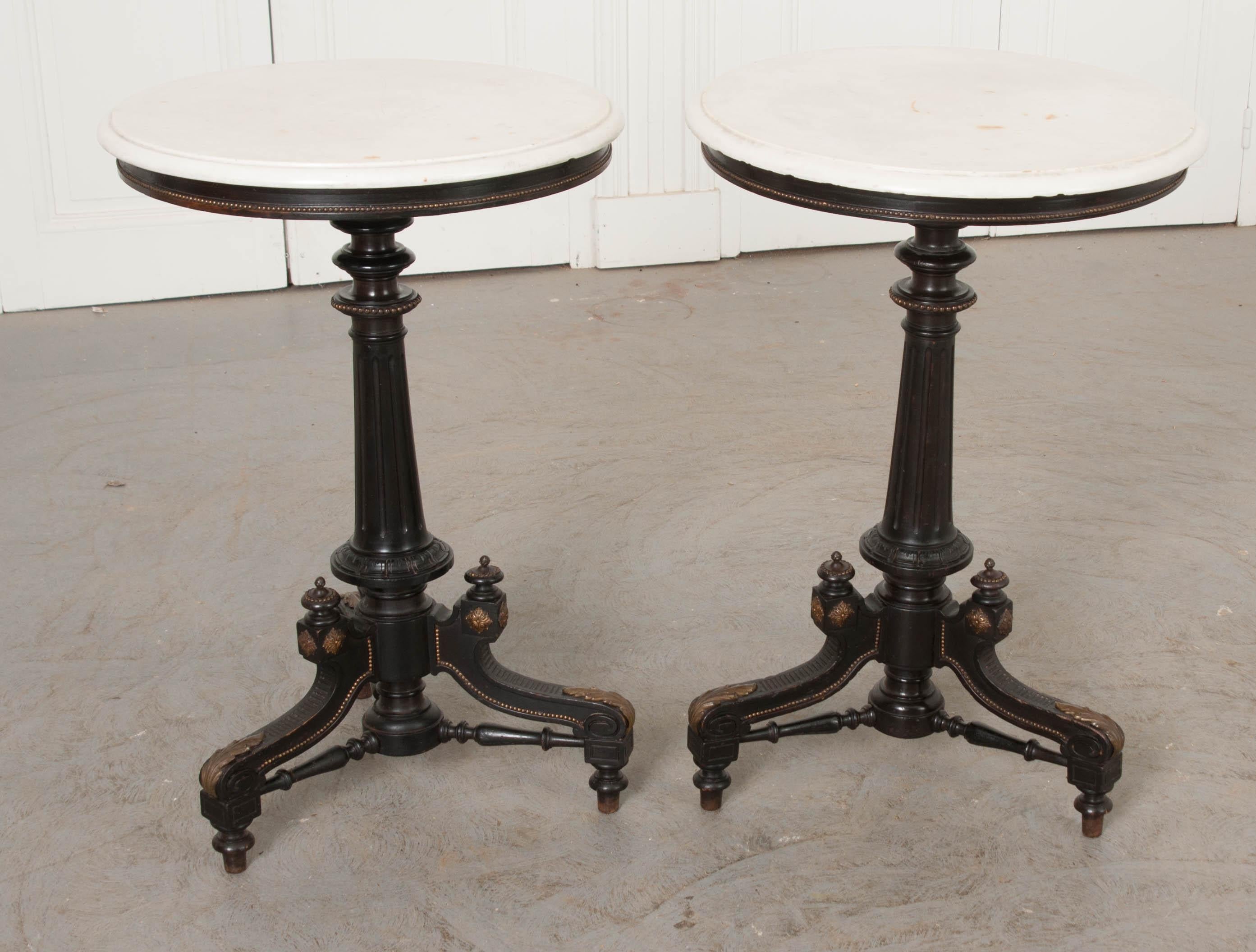 This stunning pair of ebonized marble-top tables, with impressive ormolu mountings throughout, are from Victorian England, circa 1870s. They feature carved white marble tops which screw into the beautifully turned and fluted standard above tripodal