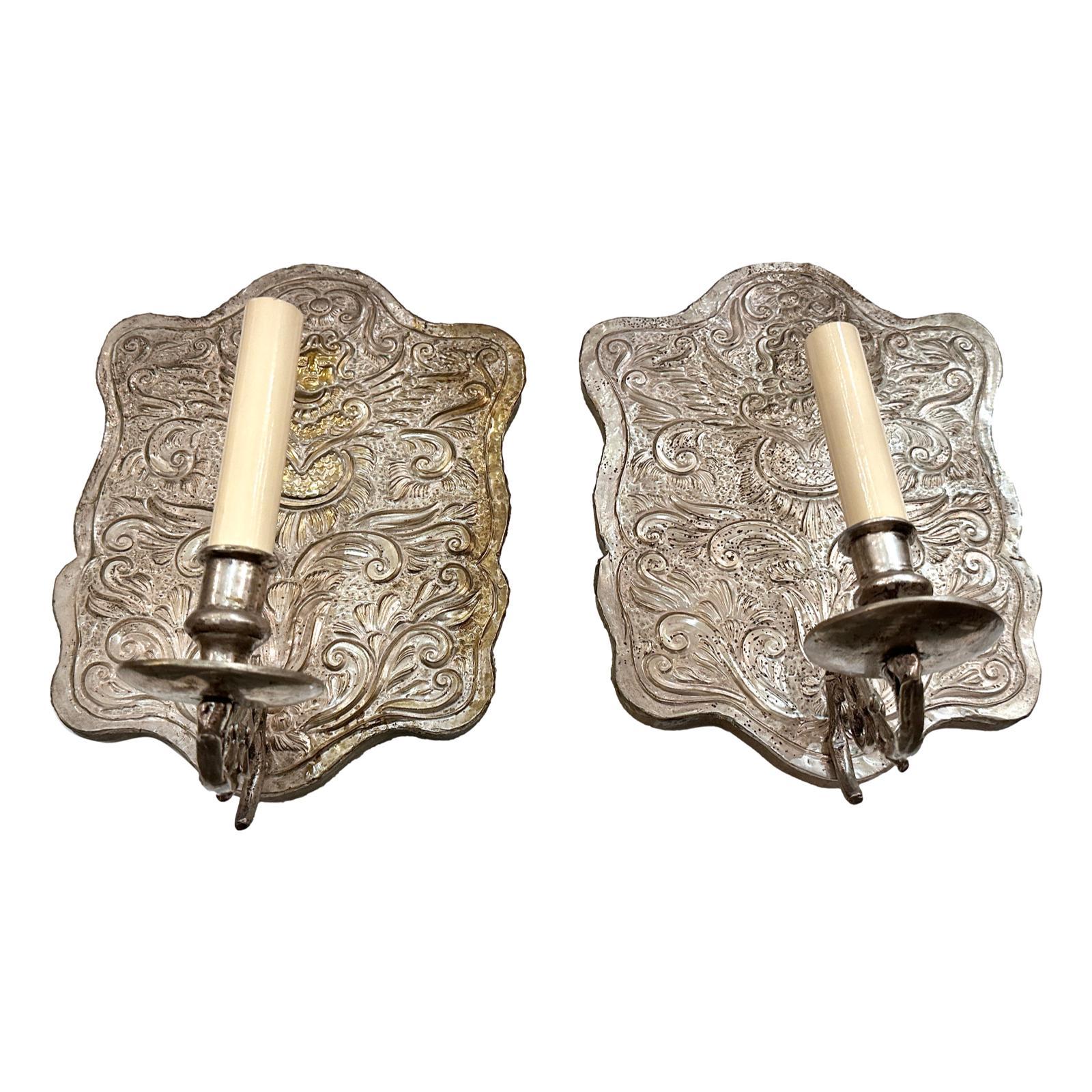 Coppia di appliques a una luce in metallo repoussé e argento placcato, risalenti agli anni '40 circa, con motivo di angeli cherubini.

Misure:
Altezza: 13,5
Larghezza: 10