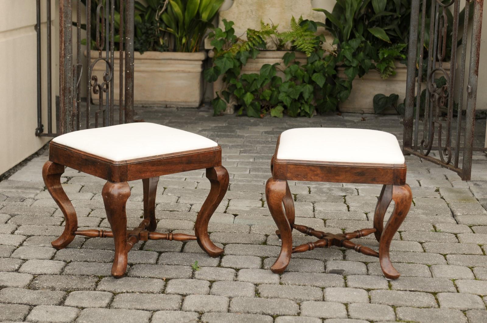 A pair of Italian Rococo style walnut stools from the mid-19th century, with cabriole legs, Saint-Andrew cross stretcher and new upholstery. Each of this pair of Italian Rococo style stools features a square seat, recovered with a simple muslin