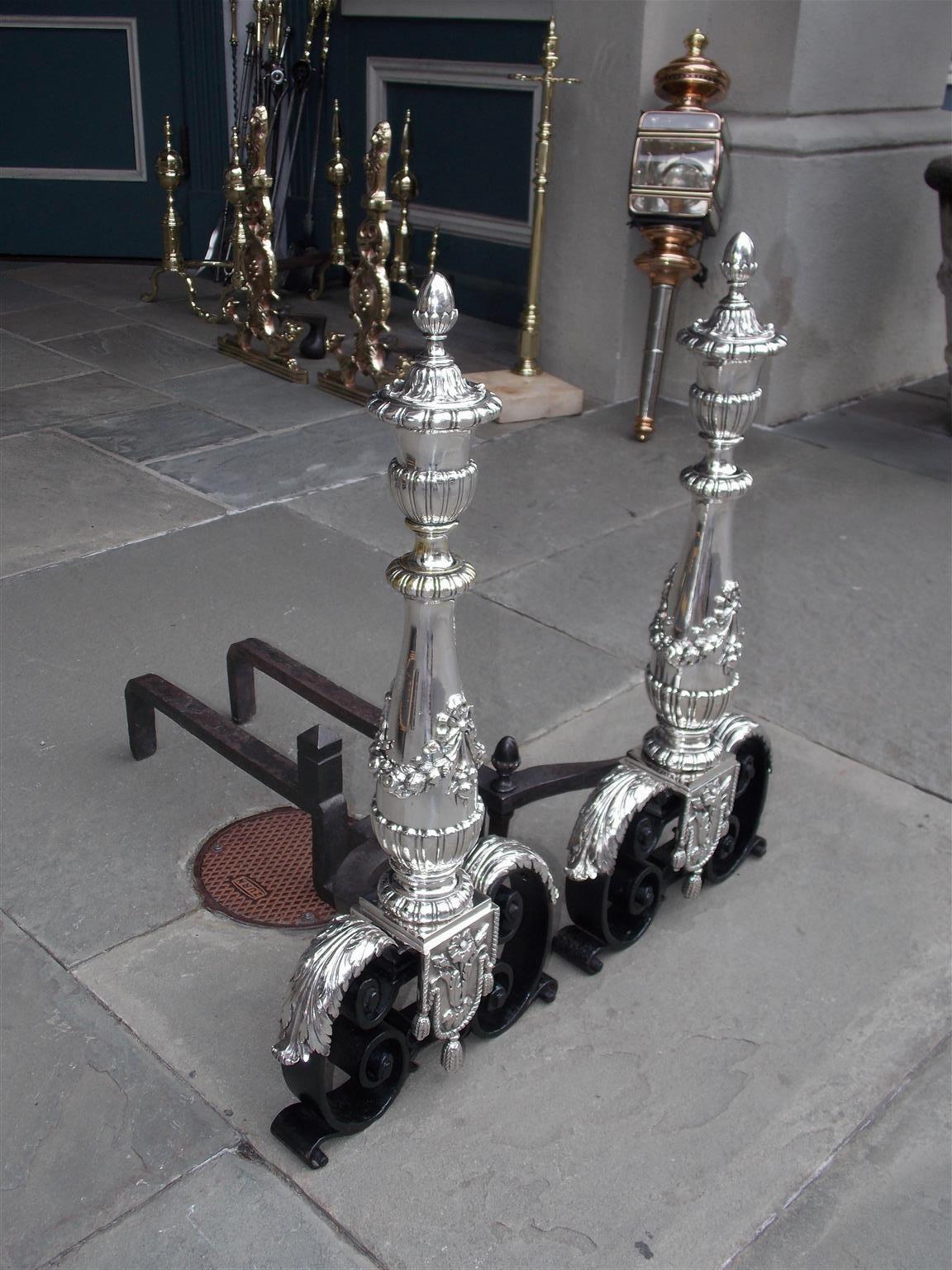 Pair of Italian silver over bronze andirons with decorative urn finials, flanking tapered columns with ribbon and floral swags, gadrooned plinths, matching finial blocked log stops, and resting on acanthus scrolled legs with floral and tassel rope