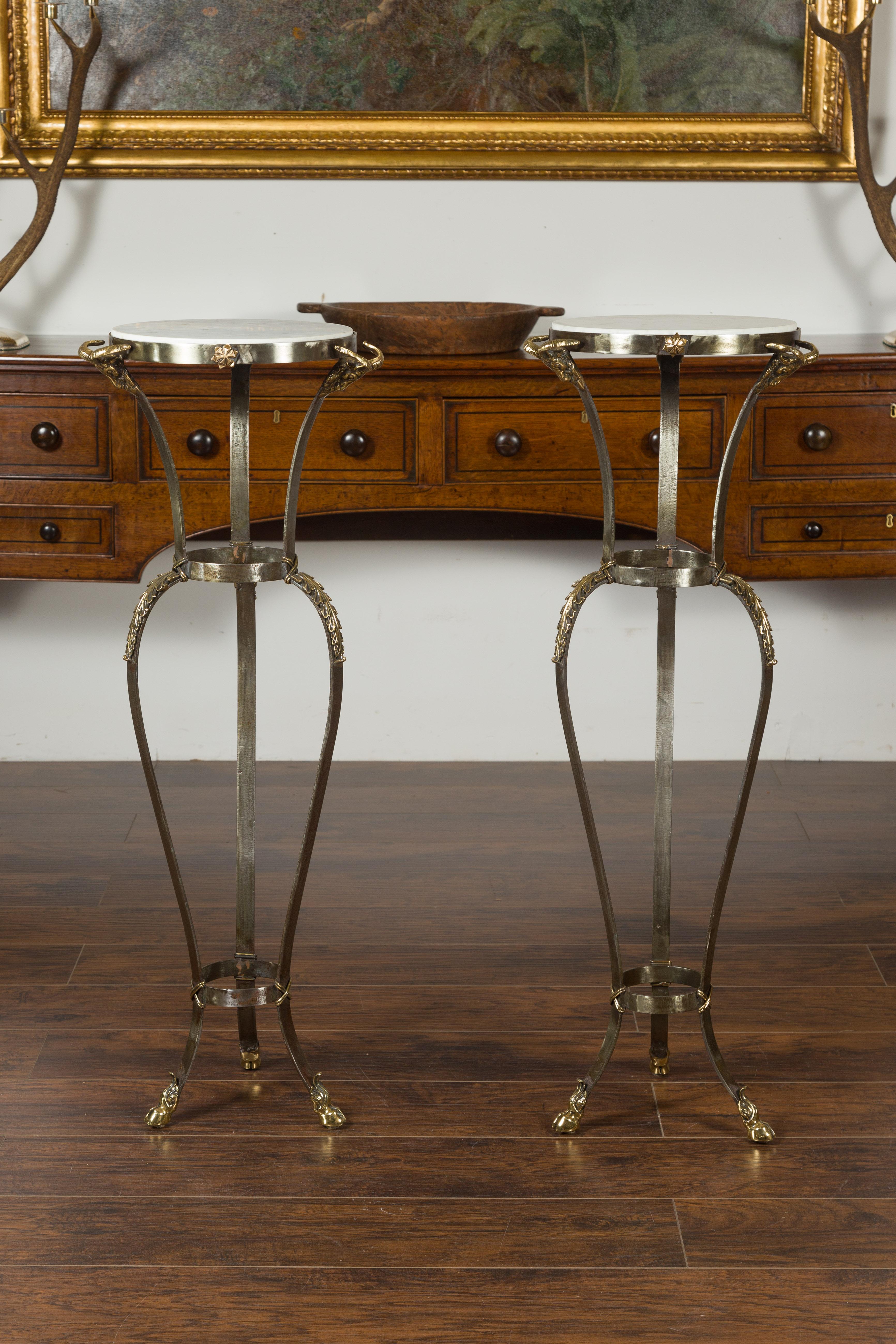 A vintage pair of Italian steel and brass pedestals from the mid-20th century with marble tops, ram's heads and hoofed feet. Born in Italy during the midcentury period, each of this pair of Italian tall pedestals features a white veined circular top