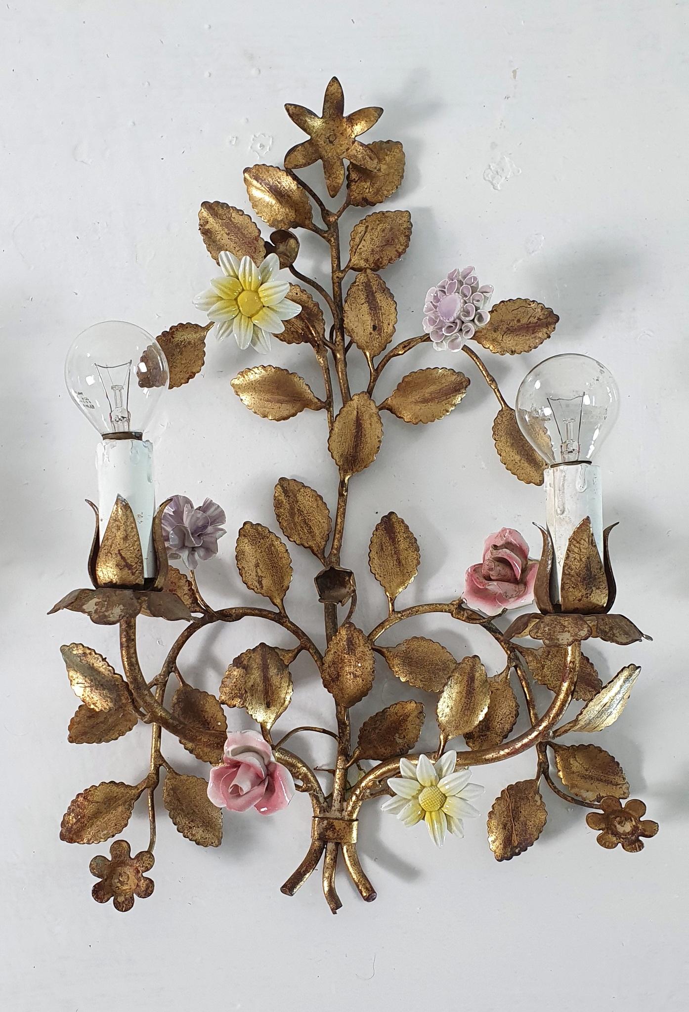 These sconces are made from iron and painted in gold color with added patina and has two candle lights each with porcelain flowers in different shapes and colors. They are both in working condition. The lightbulbs used are e14 which are found both
