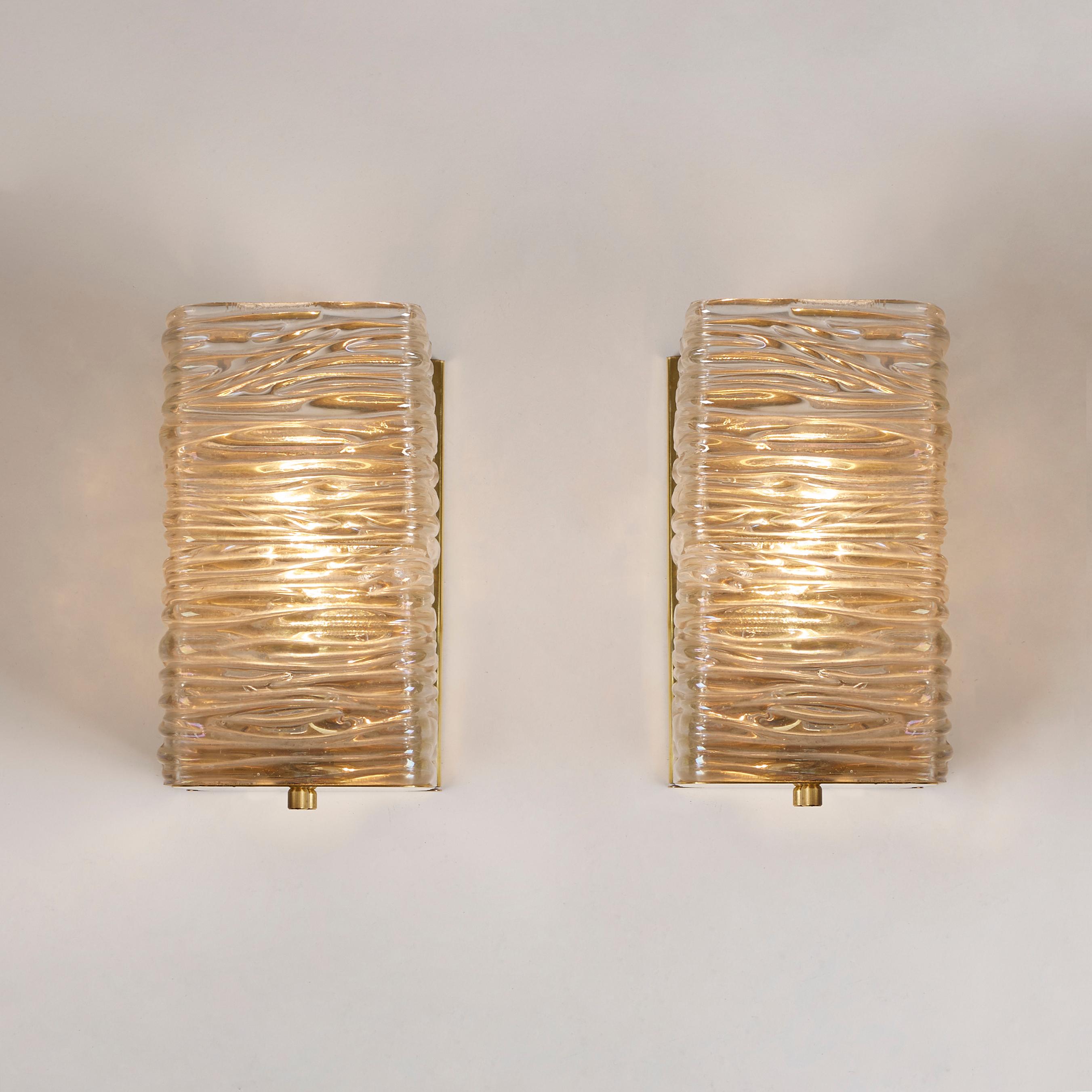 Imposantes Paar rechteckiger Wandleuchten. Zwei stark strukturierte Glaswürfel liegen auf einer Messingrückwand und einem Messingsockel. Das Glas hat eine dezent schillernde Oberfläche und strahlt ein schönes warmes Licht aus.
