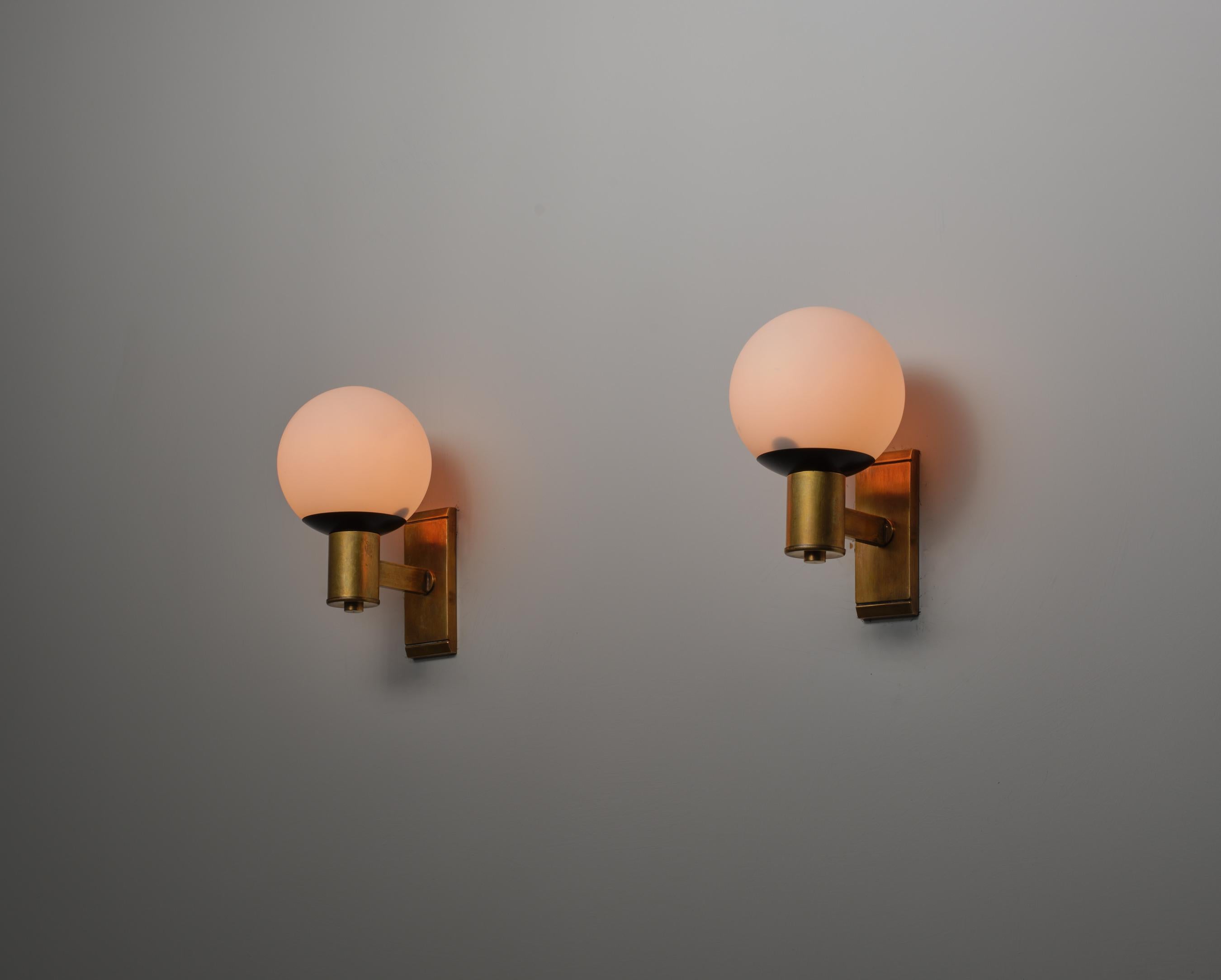 Wall-mounted sconces designed and produced in Italy during the 1950s. Each sconce features a structure composed of brass with visible patina, and a black lacquered metal base supporting a spherical opaline glass shade. The design includes a