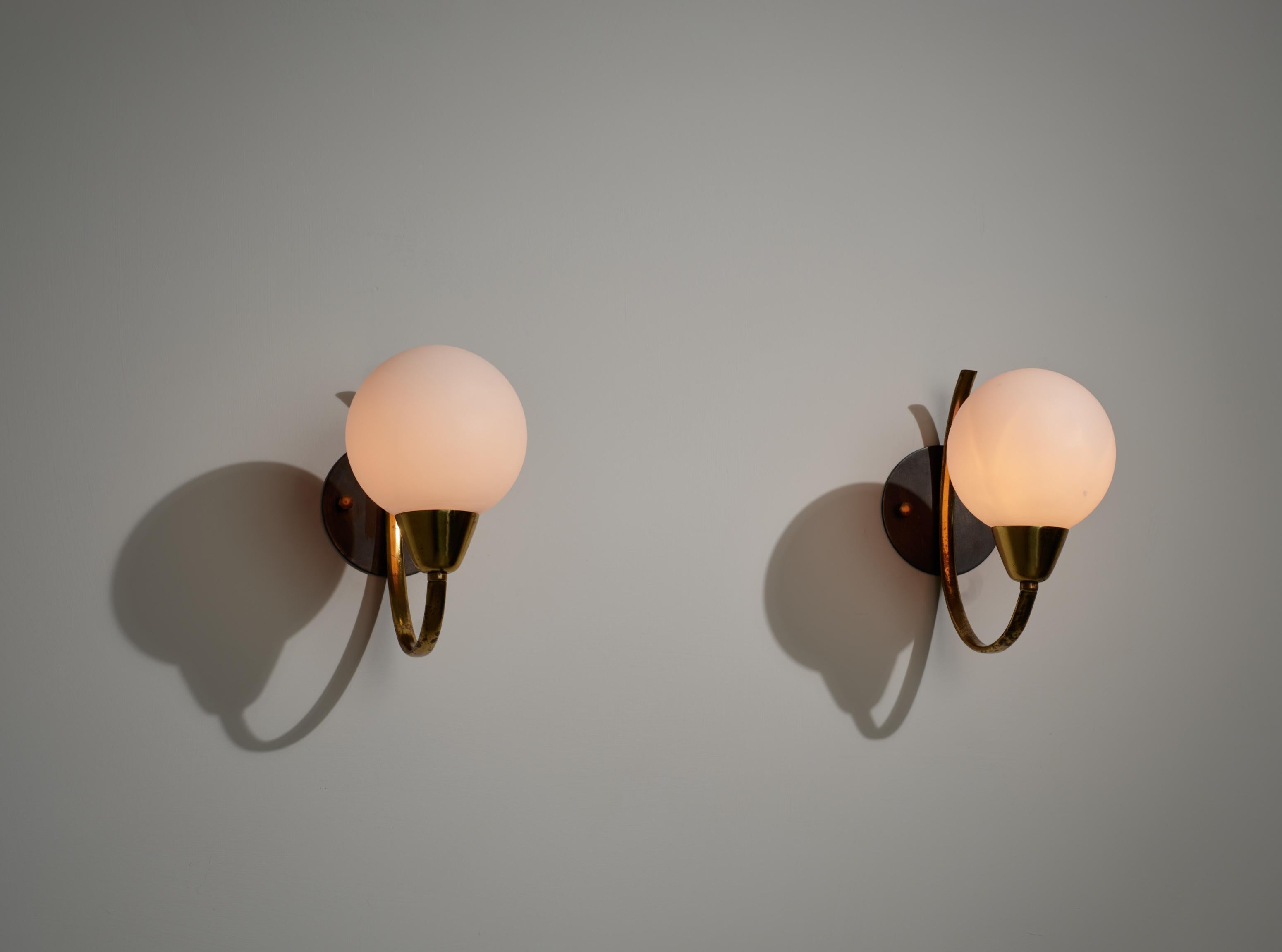 Pair of wall sconces designed and produced in Italy during the 1950s. The structure is composed of a curved brass arm with original patina, mounted on a circular backplate in burnished steel. Each sconce features a single opaline glass globe shade.