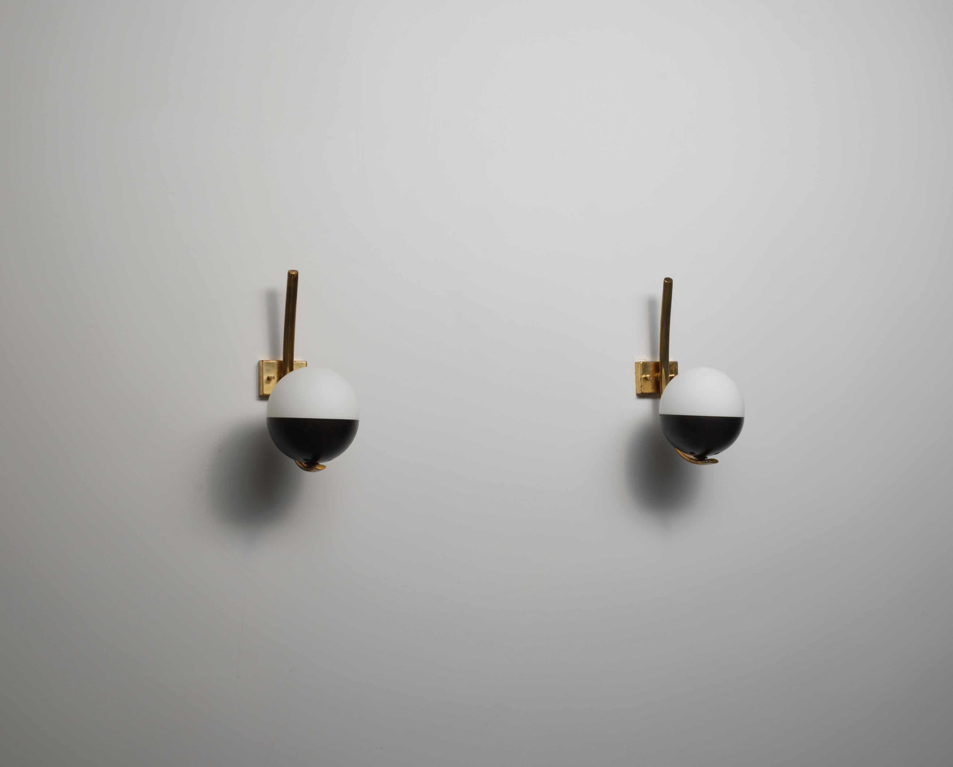 Wall-mounted sconces composed of a curved brass arm fixed to a square wall plate, supporting a spherical two-tone glass shade. The globe is made of opaline glass, with the lower half finished in burnished iron . The brass elements show a uniform