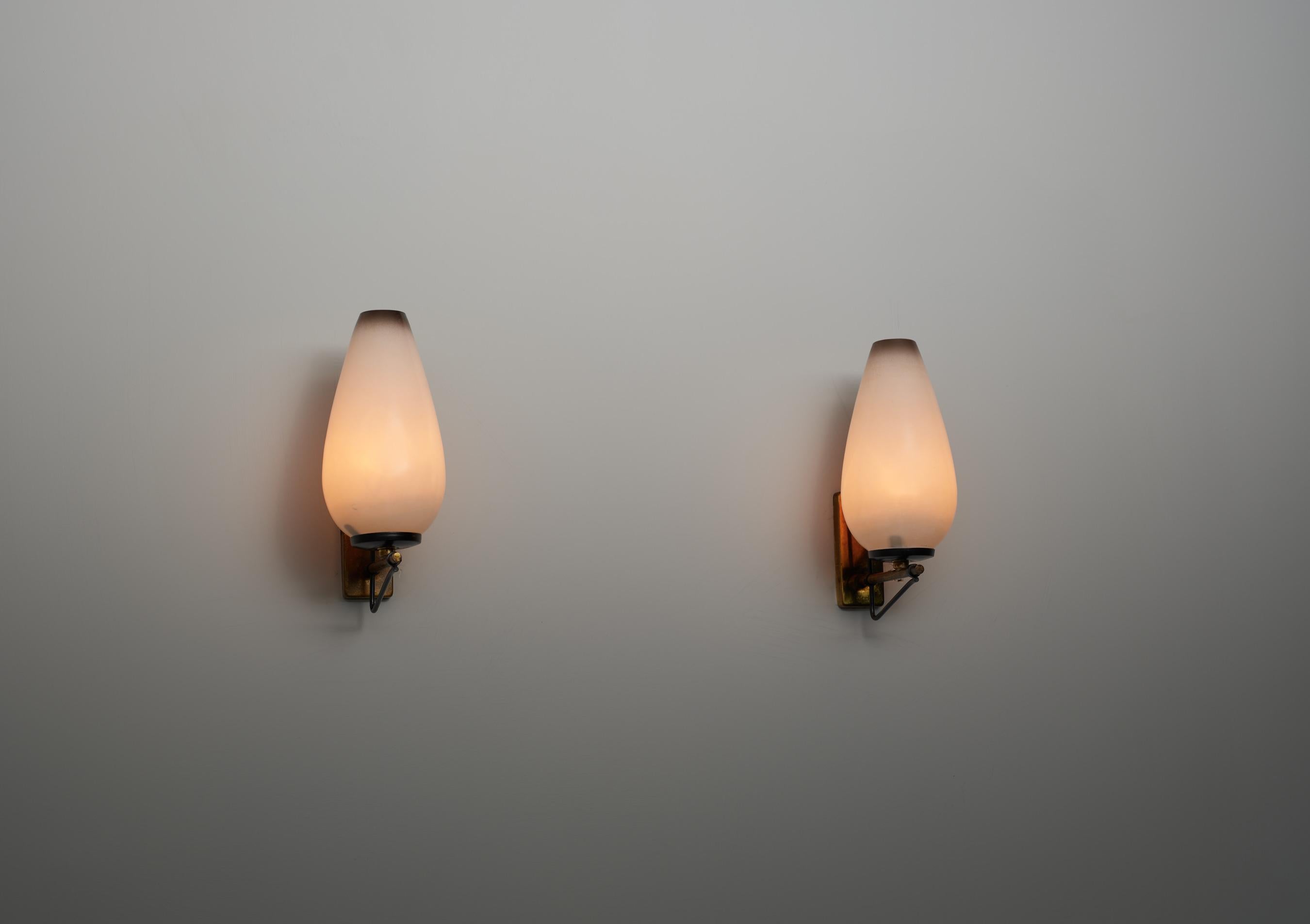 Pair of wall-mounted sconces produced in Italy during the 1950s. Each sconce features a structure in brass with a vertical wall plate and a cantilevered arm in black enameled iron. The diffuser is made of opaline glass, shaped with a subtle taper
