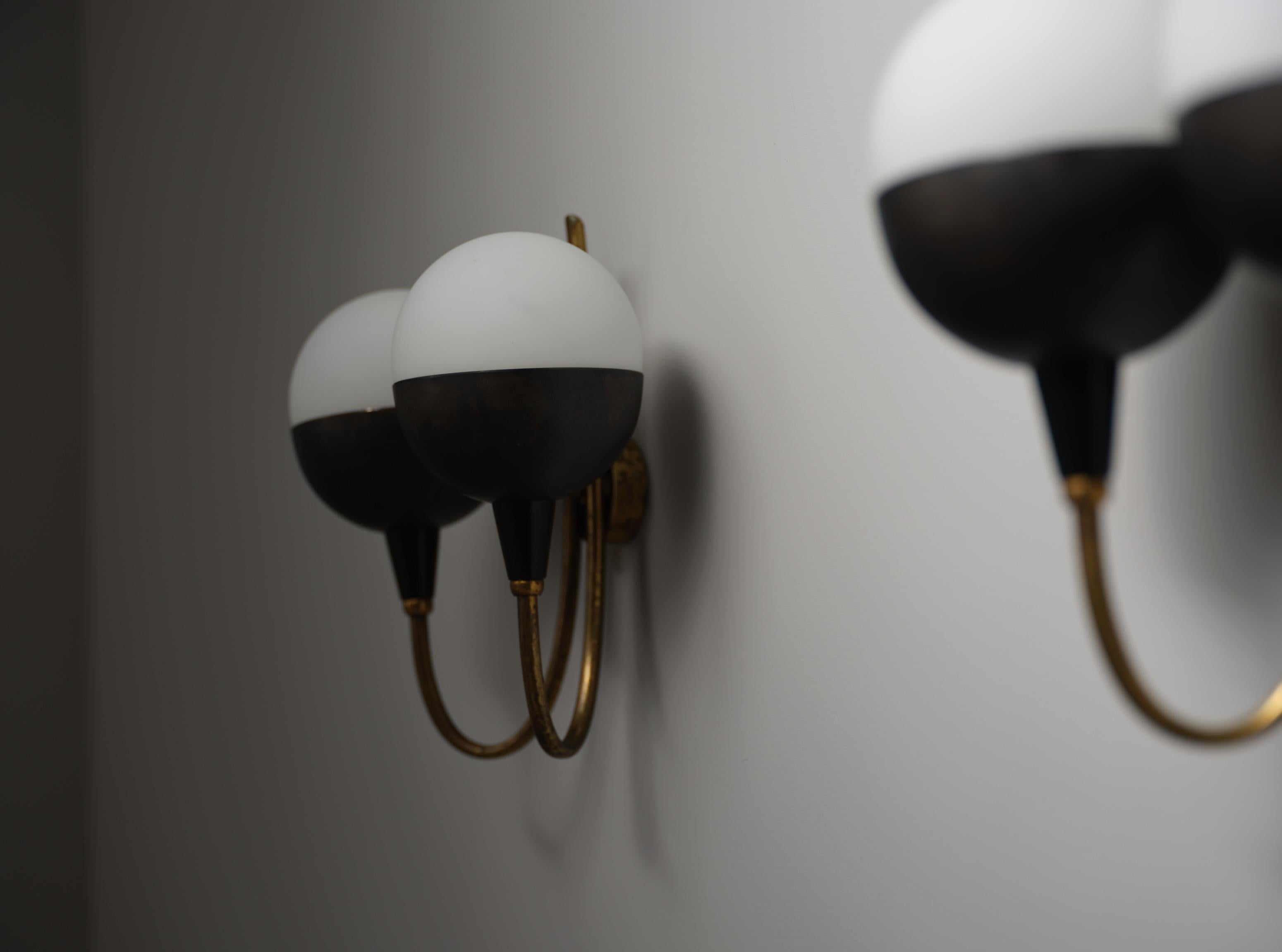 Pair of wall sconces produced in Italy during the 1950s. Each sconce features two light arms in brass with a curved, symmetrical structure, supporting spherical opaline glass shades. The globes are divided into two contrasting sections: a white
