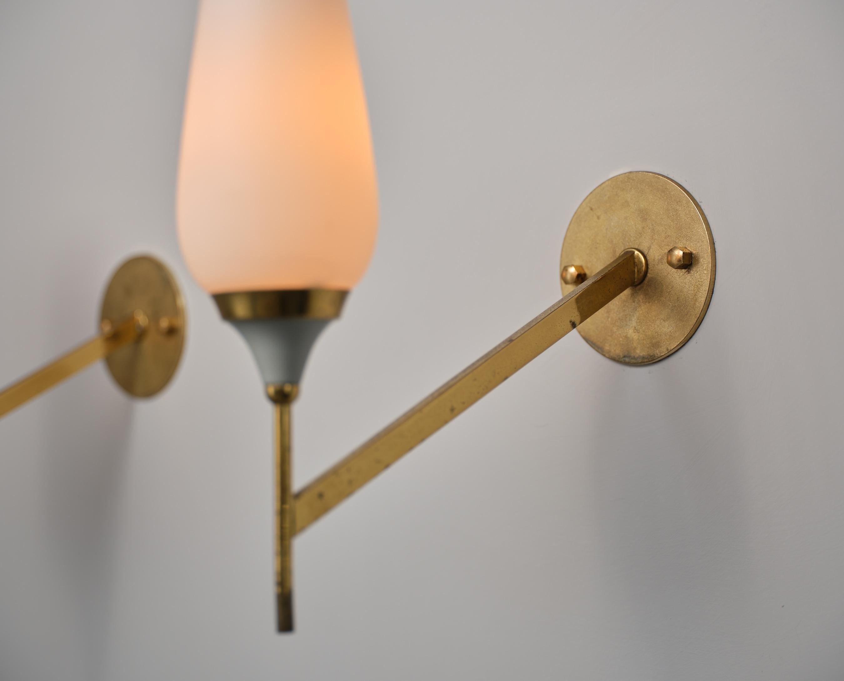 Pareja de apliques de pared diseñados y fabricados en Italia en la década de 1950. Cada luminaria tiene una pantalla cónica de cristal opalino sostenida por un brazo de latón que se extiende desde una placa trasera redonda de latón. El brazo está