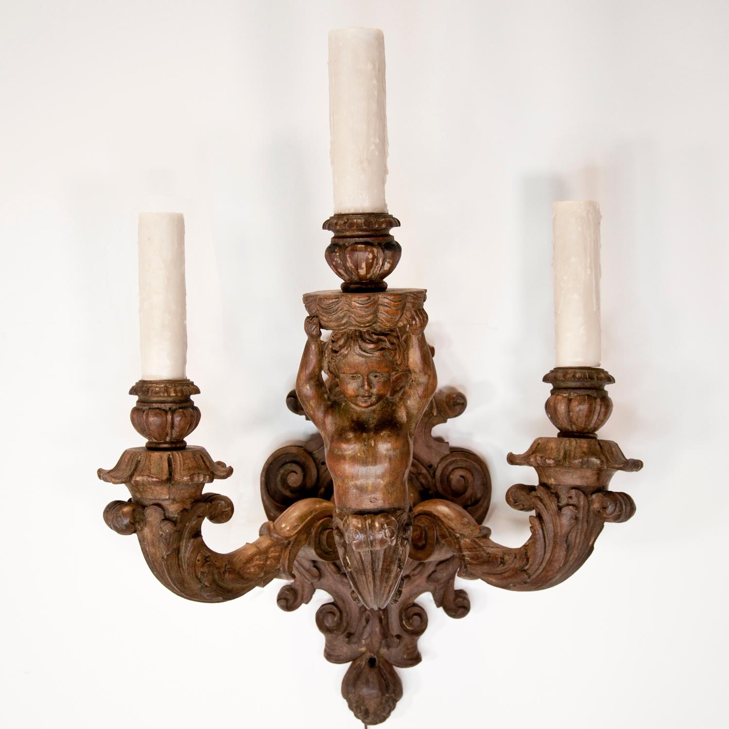 Paire d'appliques vintage en bois sculpté de style baroque, datant de la fin du 19e ou du début du 20e siècle, présentant trois bras avec des chérubins tenant un chandelier. Electrifié. Les tubes de bougies en cire au-dessus des prises de courant