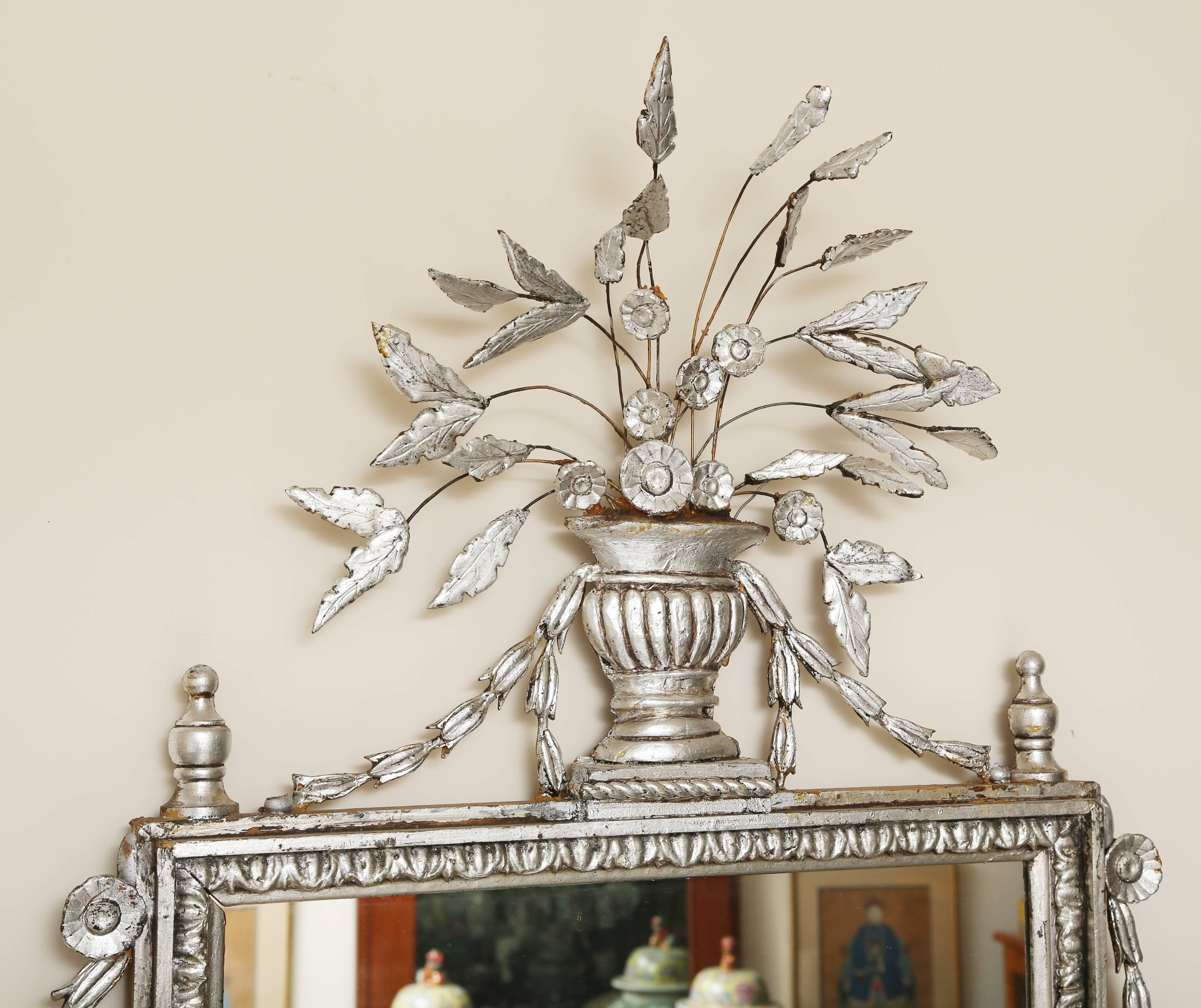 Large pair of silver leafed Italianate mirrors. The top of each mirror has an urn with an arrangement of stemmed leaves. The top, sides and bottom are decorated with swags of bellflowers. The bottom of the mirror rests on feet typical of the Italian