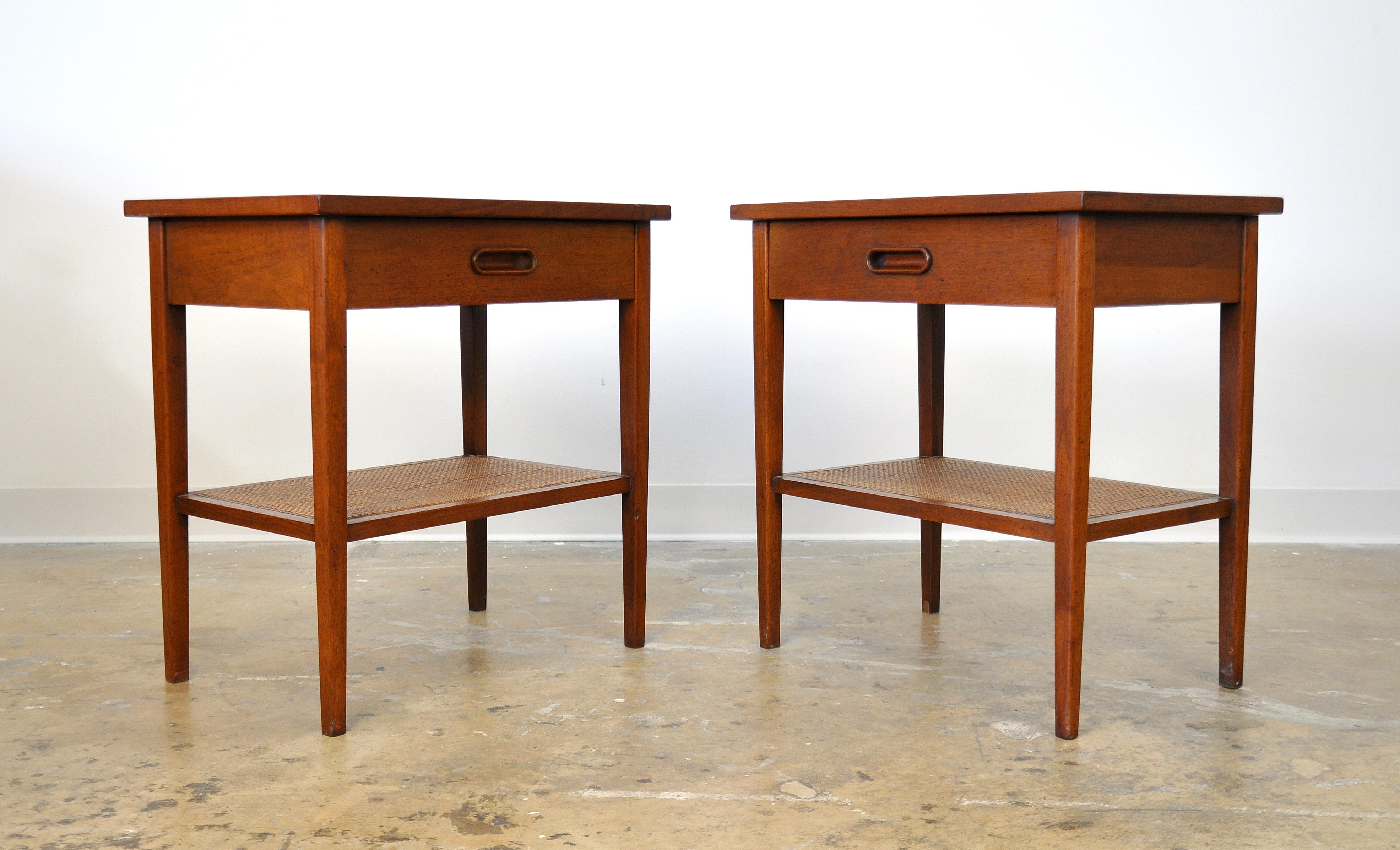 A great pair of vintage Mid-Century Modern two-tier side, end, occasional or bedside tables designed by Jack Cartwright for Founders Furniture in the 1960s. Two-tiered, fitted with a caned magazine shelf; the drawer fronts of the night stands fitted