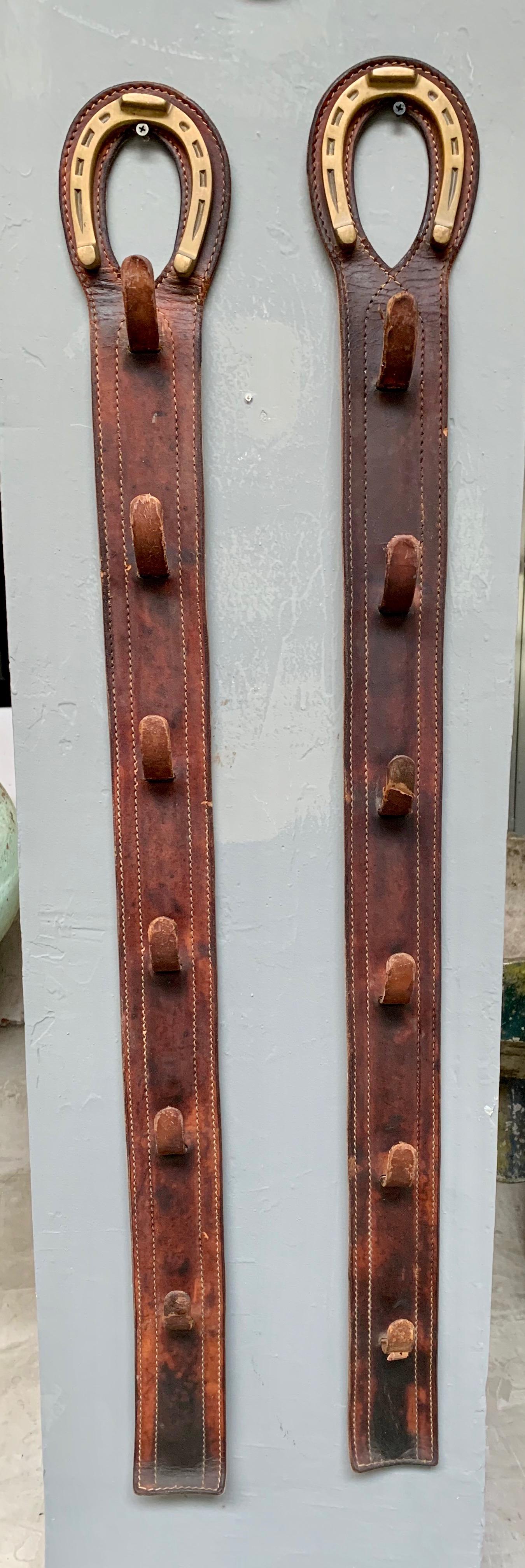 Handsome pair of coat hooks by French designer Jacques Adnet. Completely wrapped in saddle leather with brass horseshoe at top. Each pieces has a set of 6 vertical tongue hooks. Signature Adnet contrast stitching. Great patina to leather. Good