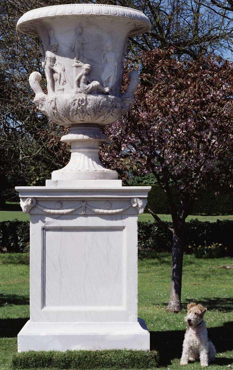 The Campana shaped urn with egg and dart and bead moulded flared rim, the body carved with the legend of Iphigenia and with fluted, leaf carved scrolled handles supported by Bacchic masks, the turned and fluted socle with a carved collar. Rome’s