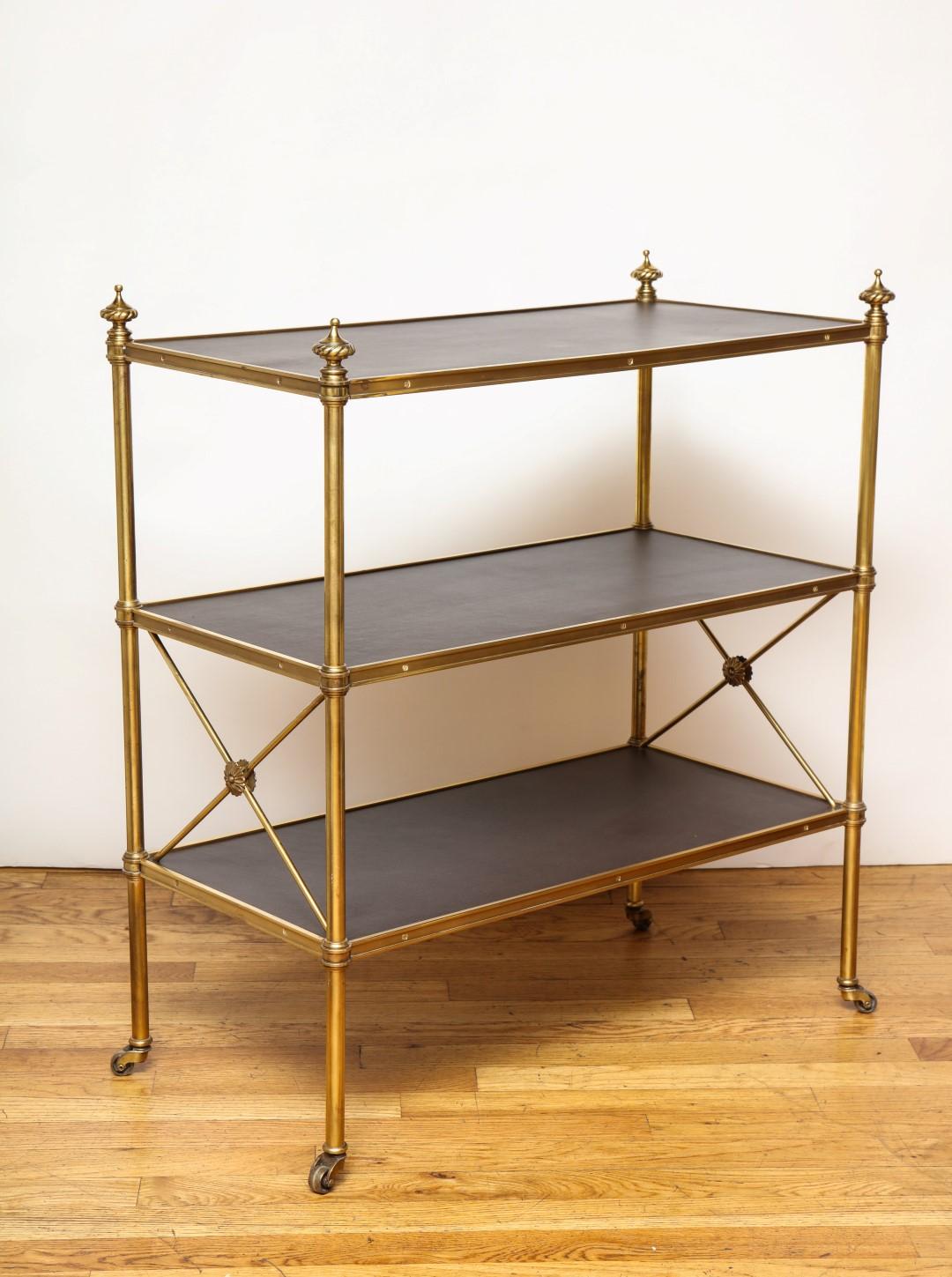 A pair of Jansen style brass etagères, the frames with rectangular leather covered wooden surfaces and x-shaped supports with centre rosettes on lower tier. The legs issuing small casters.
  