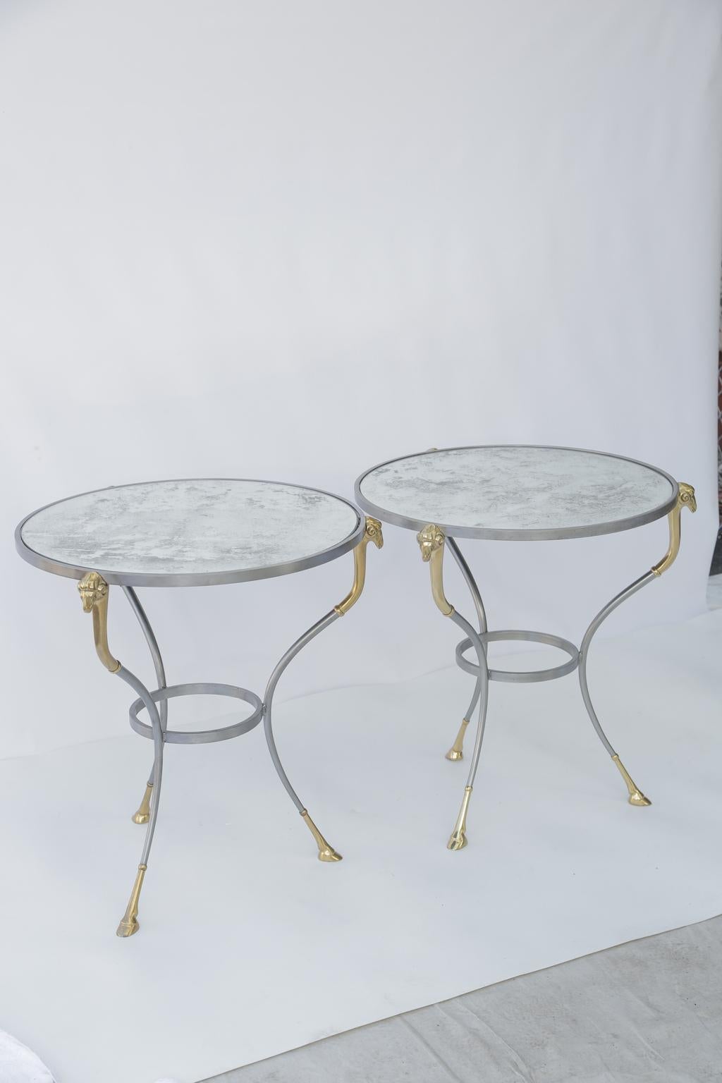 Pair of gueridon end tables, in the manner of Maison Jansen, each having a round top of antiqued mirror, on table bases of polished steel, raised on three curving legs, headed with ram's masks of brass, ending in cloven-hoofed feet, and joined by a