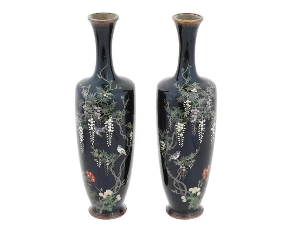 A matched pair of antique Japanese vases of elongated shape with long flared necks. Meiji period, 1868-1913. The pieces are decorated with silver wired polychrome cloisonne enamel scenes depicting birds perching among branches of wisteria flowers