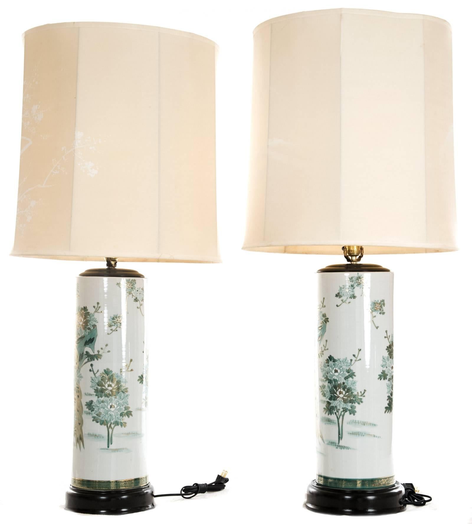 A pair of Japanese Kutani porcelain table lamps of cylindrical form with hand-painted decoration in tones of green with gold-enhanced details and highlights depicting peacocks amongst lush florals, mounted on an ebonized wood base. The tall drum