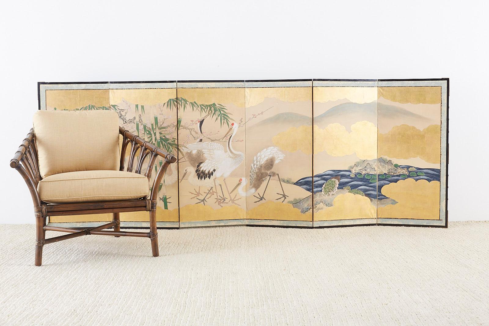 Fascinating pair of Japanese six-panel Meiji period screens depicting red-crowned cranes and minogame in landscape scenes. The colorful landscapes feature pine, bamboo, camellia flowers, and a blue seashore on one screen. The screens are titled on