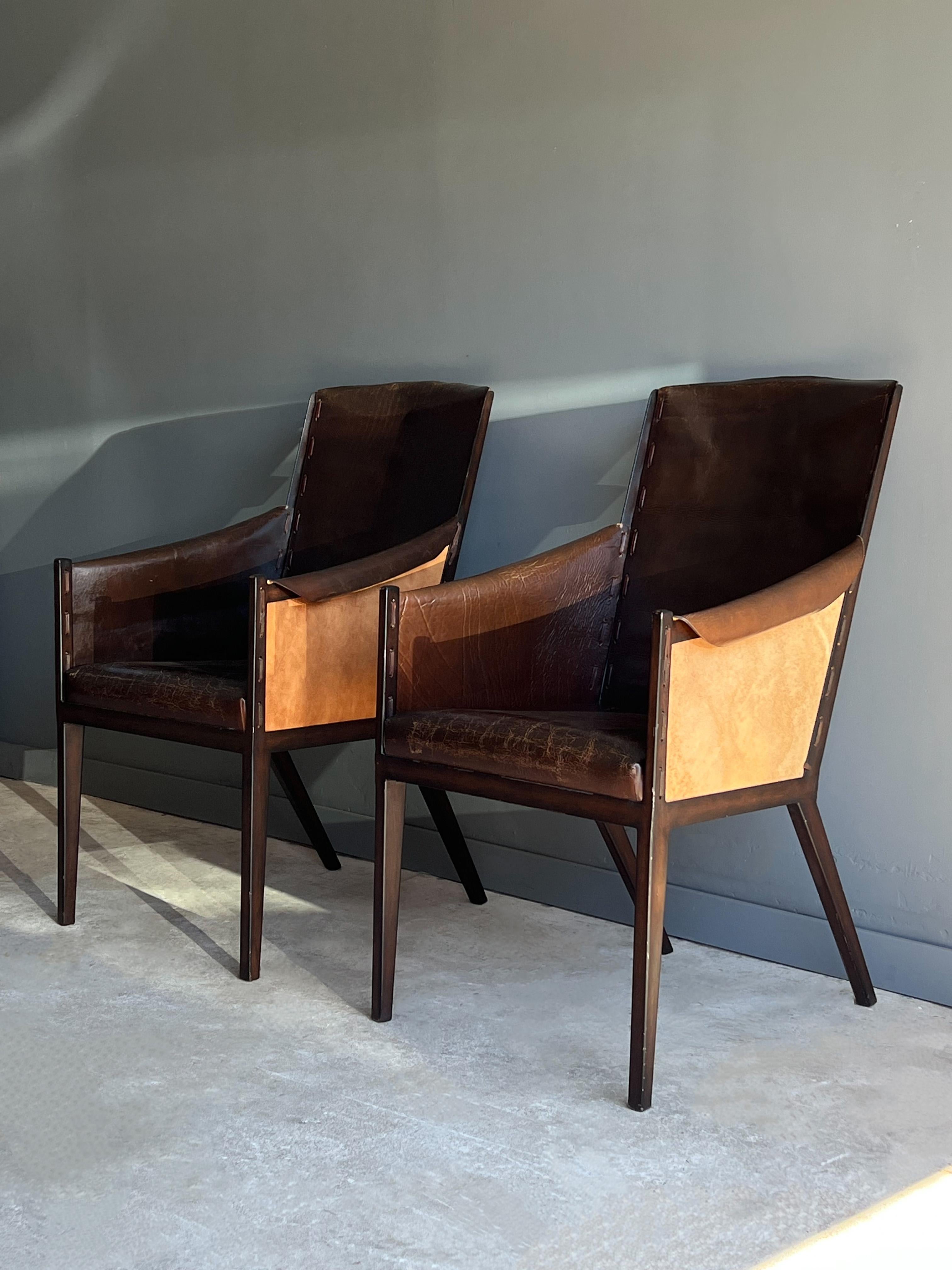 A lovely pair of French style arm chairs in the manner of Jean Michel Frank. Each chair is made with a thick leather and heavy gauge metal frames. The leather shows a nice patina and weathered appearance. Sides and back are whipped stitched onto the
