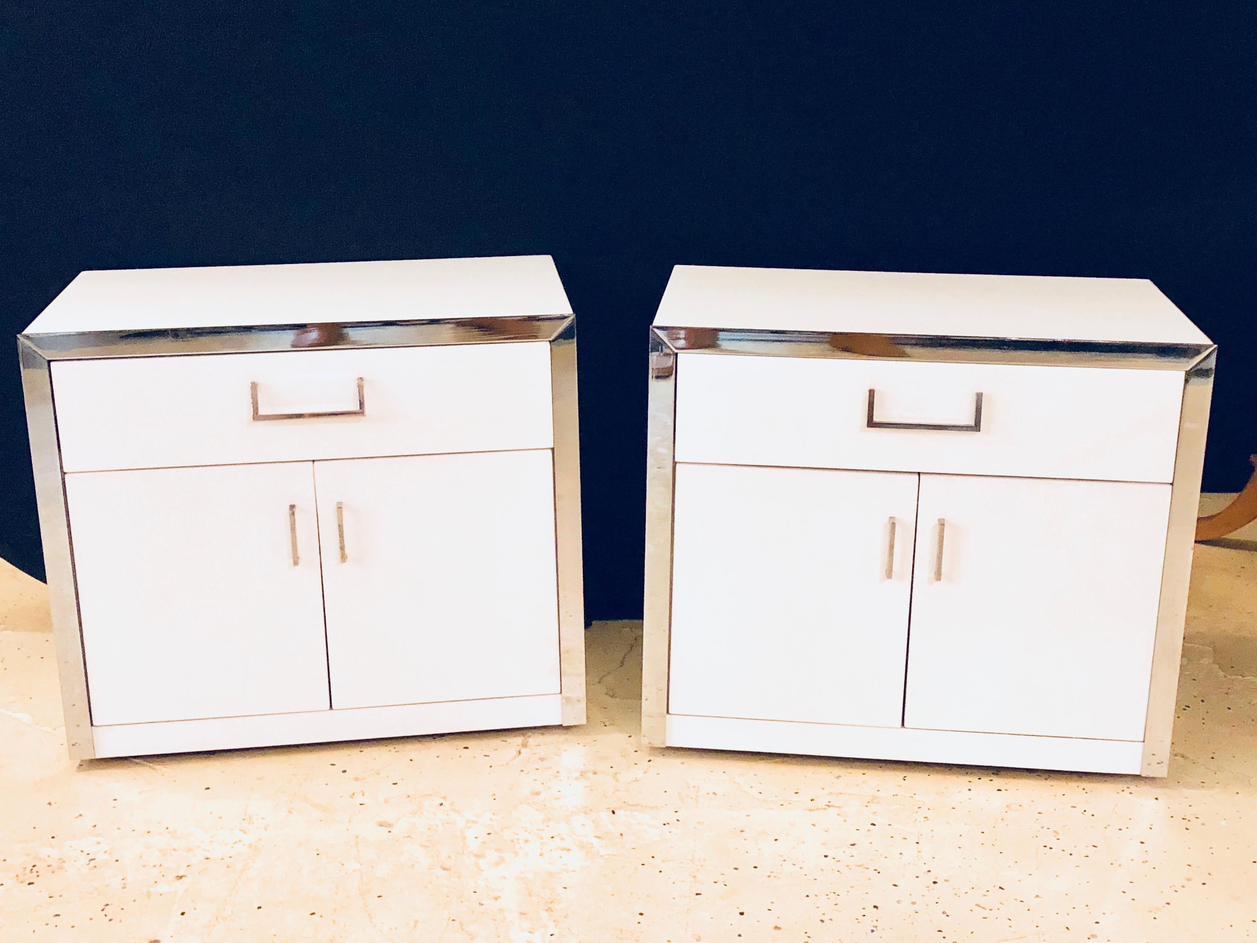 Pair of labeled John Stuart Mid-Century Modern white and chrome decorated nightstands or end tables. Each having two doors leading to a storage area under a single drawer. The doors and drawers with chrome pulls and a chrome frame around the front.
