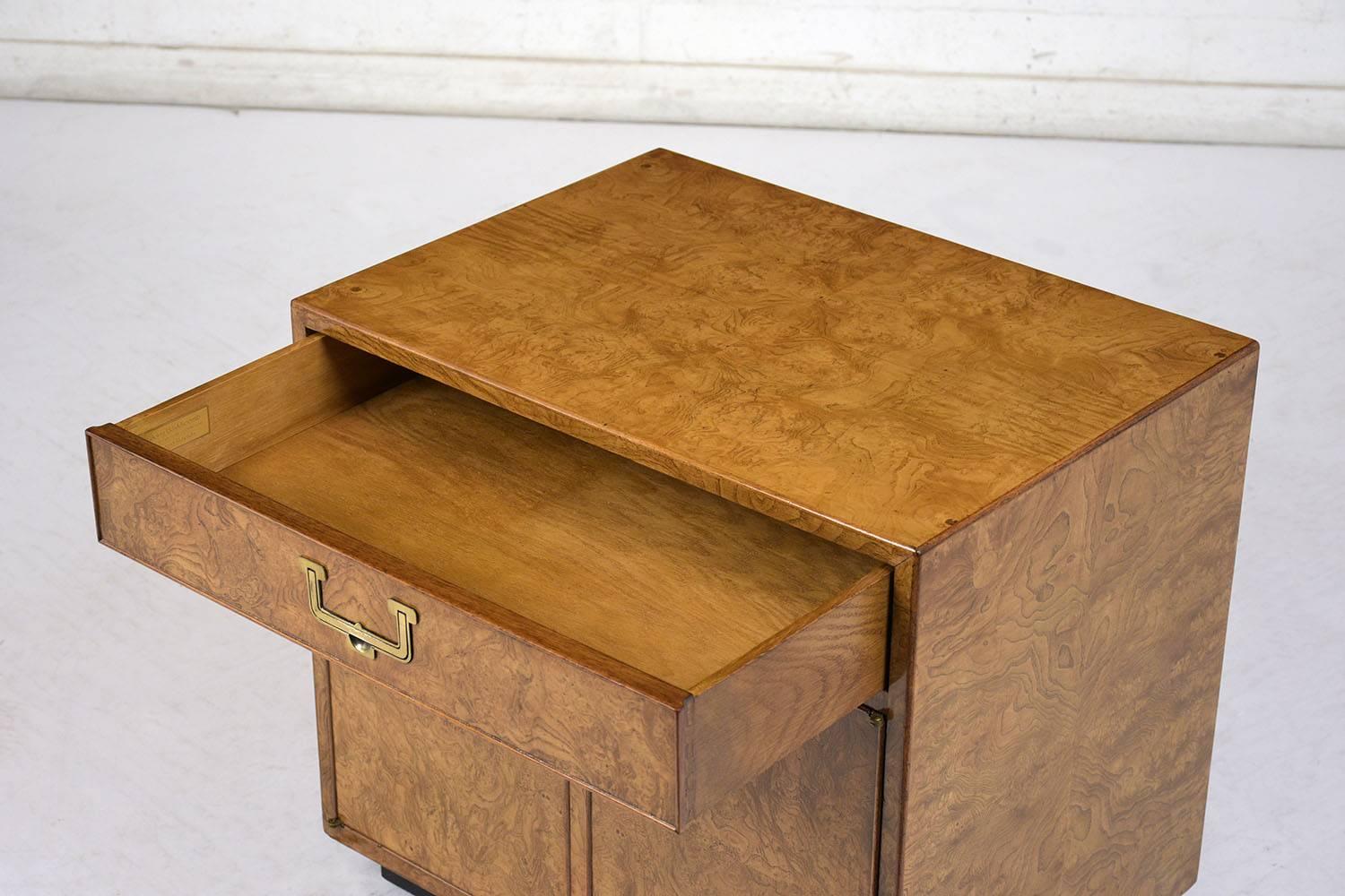 This pair of 1960s Campaign-style nightstands are made by John Widdicomb. The nightstands are adorned with burled wood veneers finished in a walnut color with black square feet. There is a single drawer and a two door cabinet below for ample storage