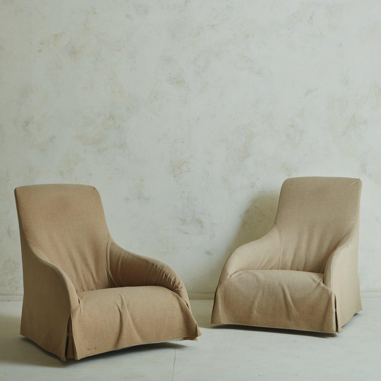 A pair of ‘Kalos’ swivel lounge chairs designed by Antonio Citterio and manufactured by Maxalto in Italy. These sculptural chairs feature tall, angled seat backs and curved arms. They retain their original woven tan slipcovers. Retain ‘B&B Italia /