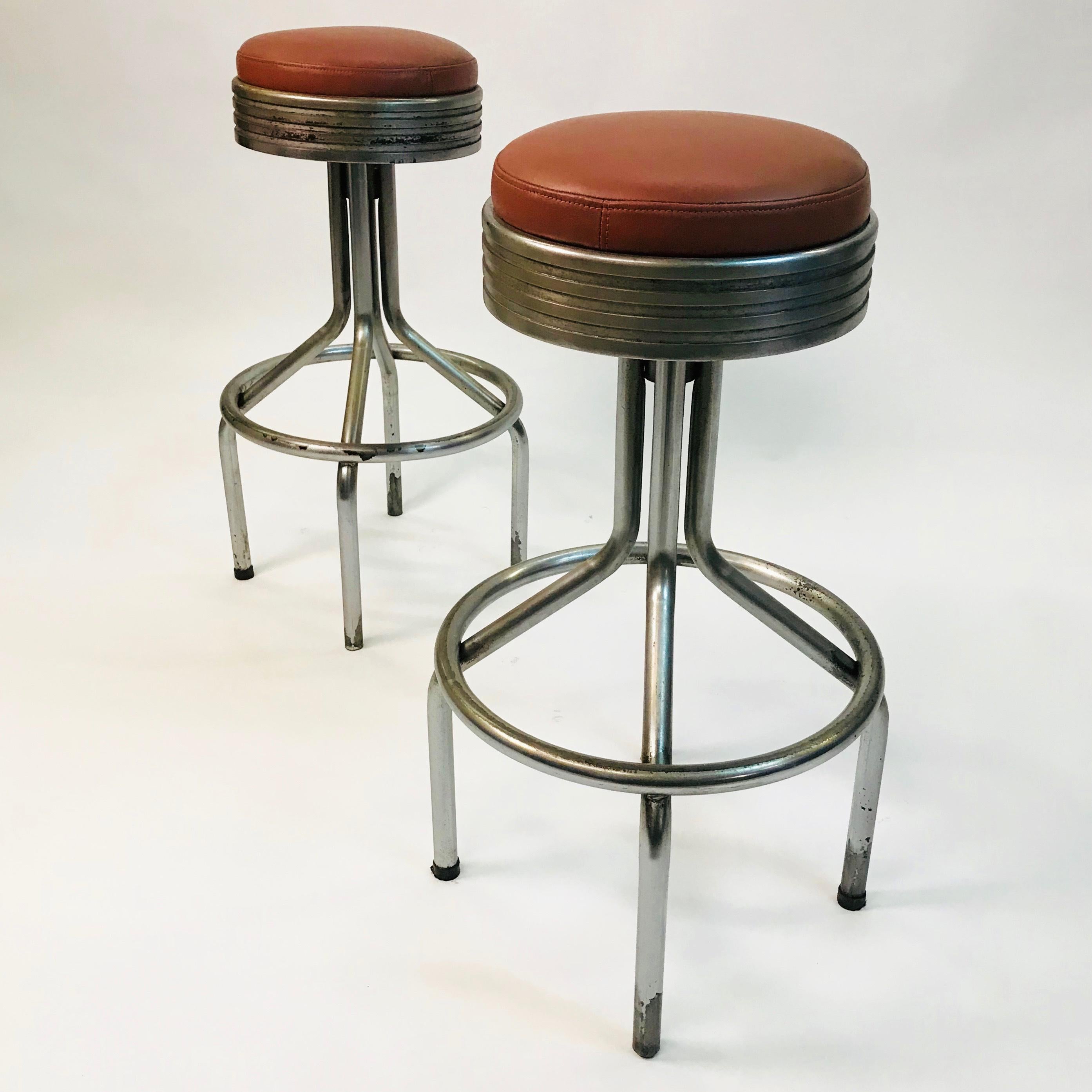 Pair of Machine Age, Art Deco, bar stools by KEM Weber feature tubular chrome bases with 17.5 inch diameter foot rests and newly upholstered, chrome-plated cast iron, swivel, leather seats that are 13.5 inch diameter. The footrests are at 12.5