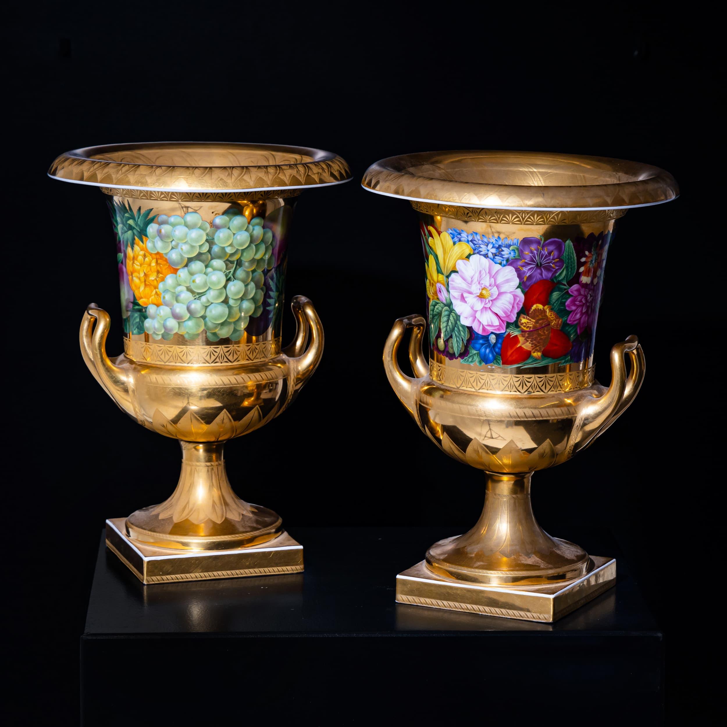 Coppia di vasi a cratere in porcellana con fondo dorato di forma classica. I corpi sono dipinti con colori vivaci: uno con frutti come ananas, uva e mele; l'altro con fiori esotici, in particolare una sorprendente Strelitzia. Entrambi i vasi sono in