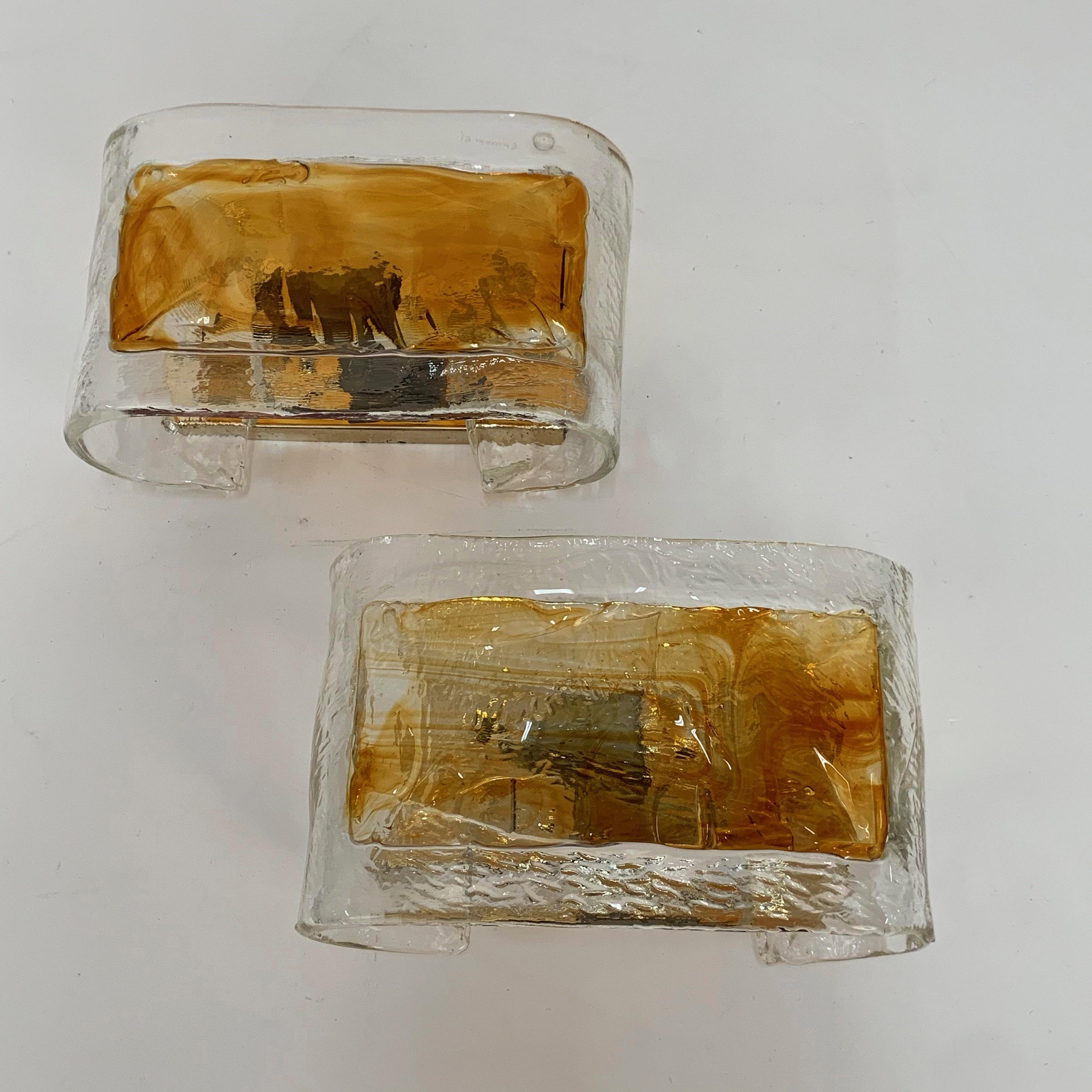 Pair of amber Murano glass wall lights. Produced by La Murrina in Italy during the 1970s.

They consist of two elements, an amber coloured Murano glass element resting on a golden metal base. Both elements influence the refraction of light,